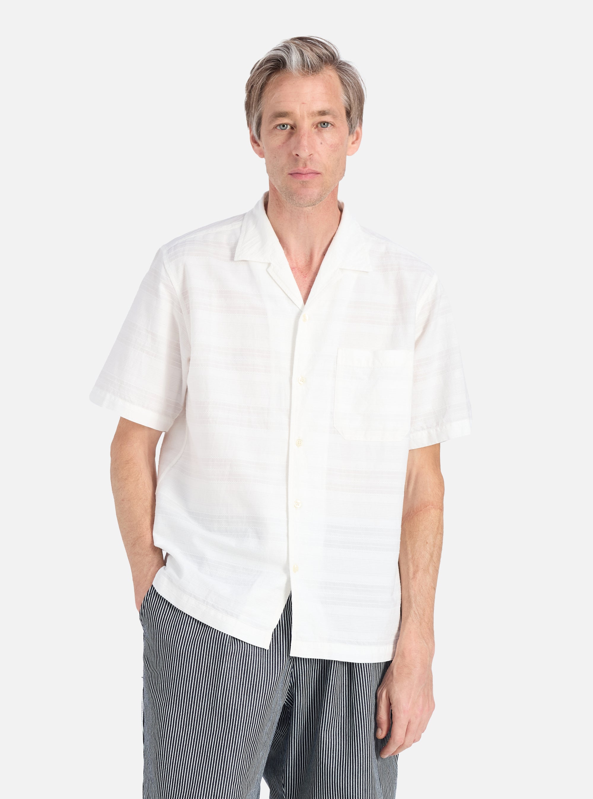 A model on a white background wears the Ecru Deffe Self Stripe Minari Shirt. The shirt is a light color with subtle stripes and a relaxed fit.