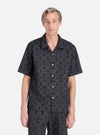 A model stands on a white background wearing the Navy Mitla Print Cotton Road Shirt. The shirt is navy and brown with a geometric print and has short sleeves.
