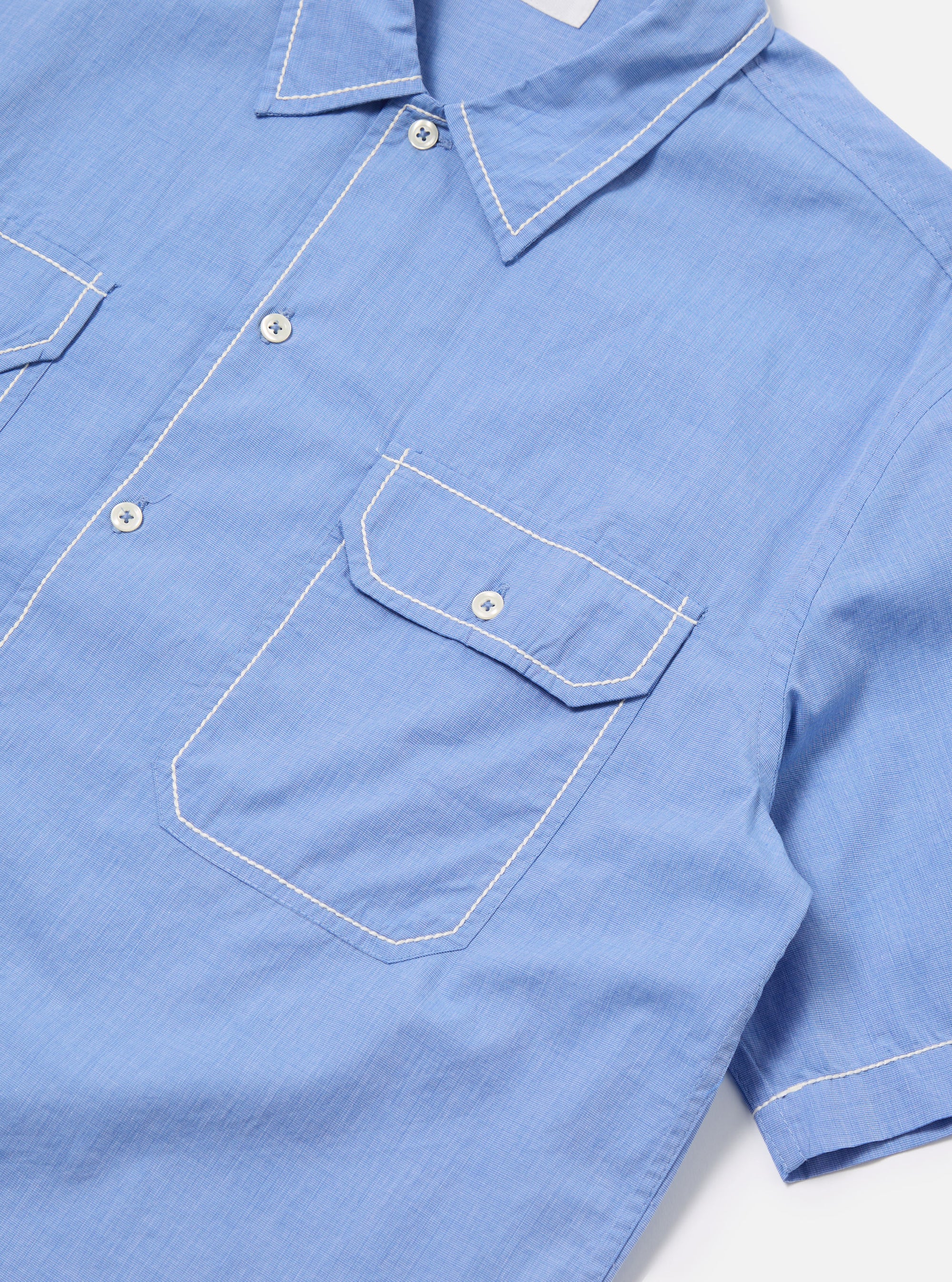 Close-up of the Blue End On End Cotton Utility SS Shirt on a white surface. The light blue shirt features short sleeves, white buttons, and two chest pockets with white stitching.