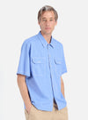A man on a white background wears the Blue End On End Cotton Utility SS Shirt. The shirt is light blue with white stitching and two chest pockets. He also wears tan pants.