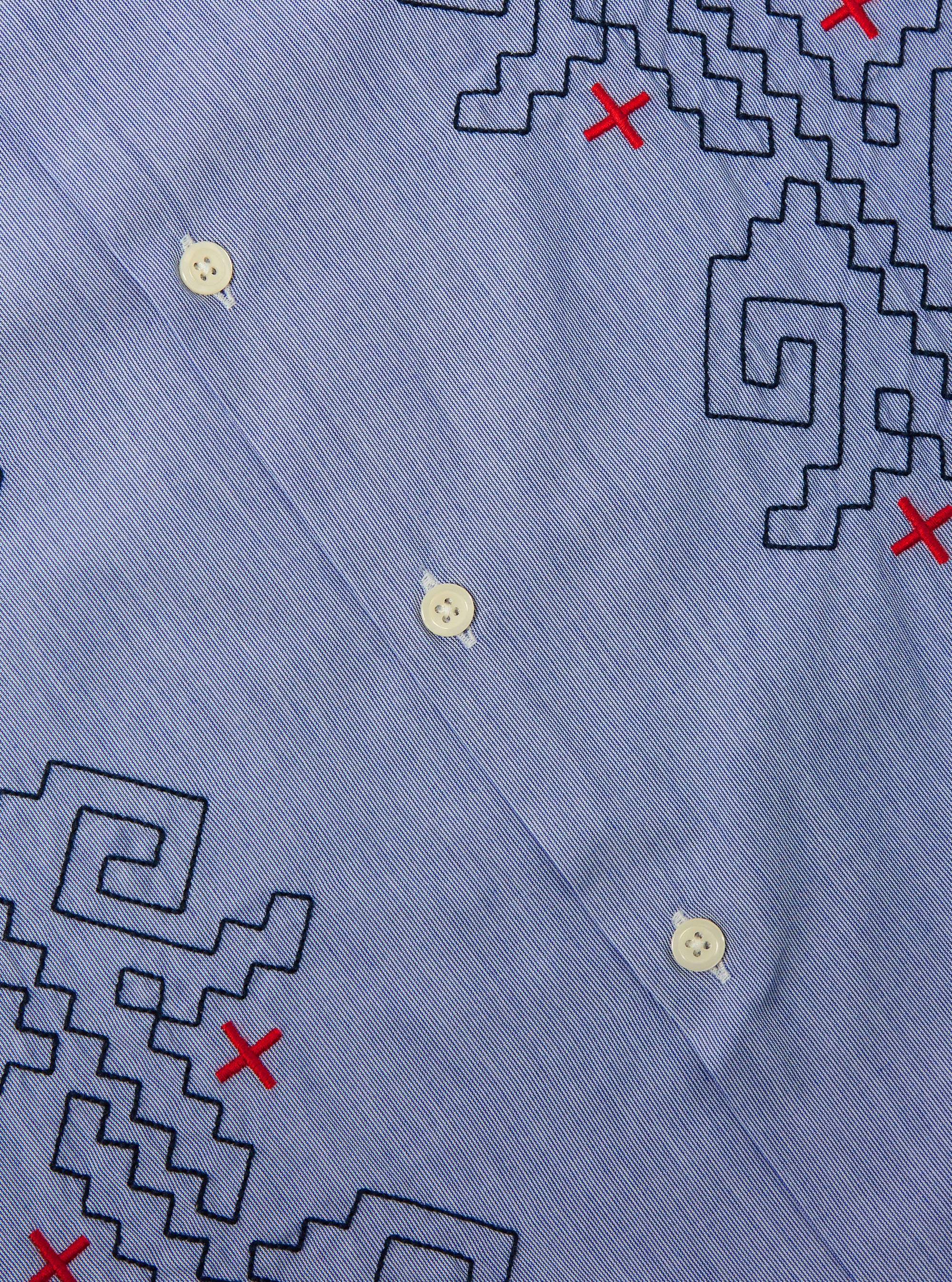 Close-up of the Blue Classic Stripes Road Trip Shirt. Light blue fabric with black geometric embroidery and red crosses. White buttons are visible.