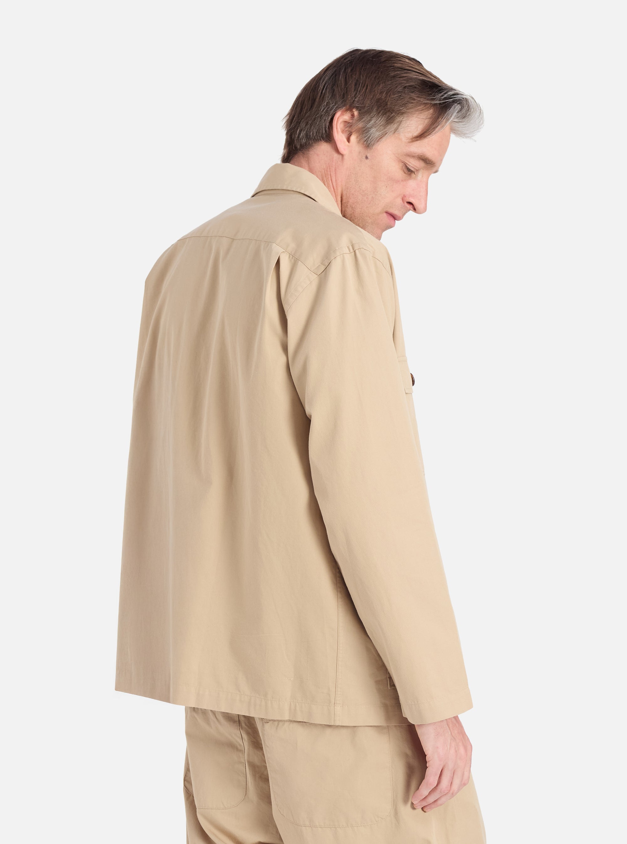 A man models the Sand Military Cotton Dockside Overshirt in a studio setting. The overshirt is a light sand color and appears to be made of cotton. The background is a clean white.