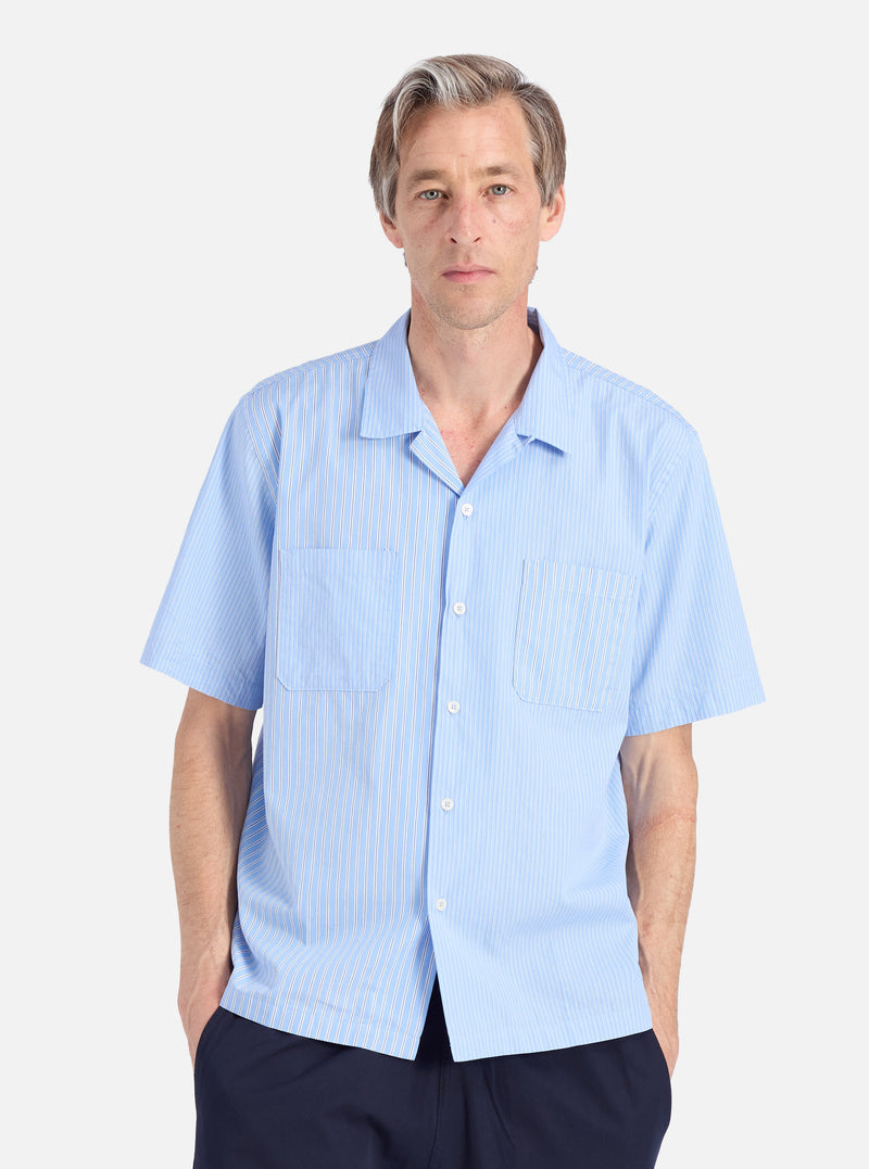 A man models the Blue Vila Stripes Sal Road Shirt. The light blue shirt has white stripes and short sleeves. Shot against a white backdrop.