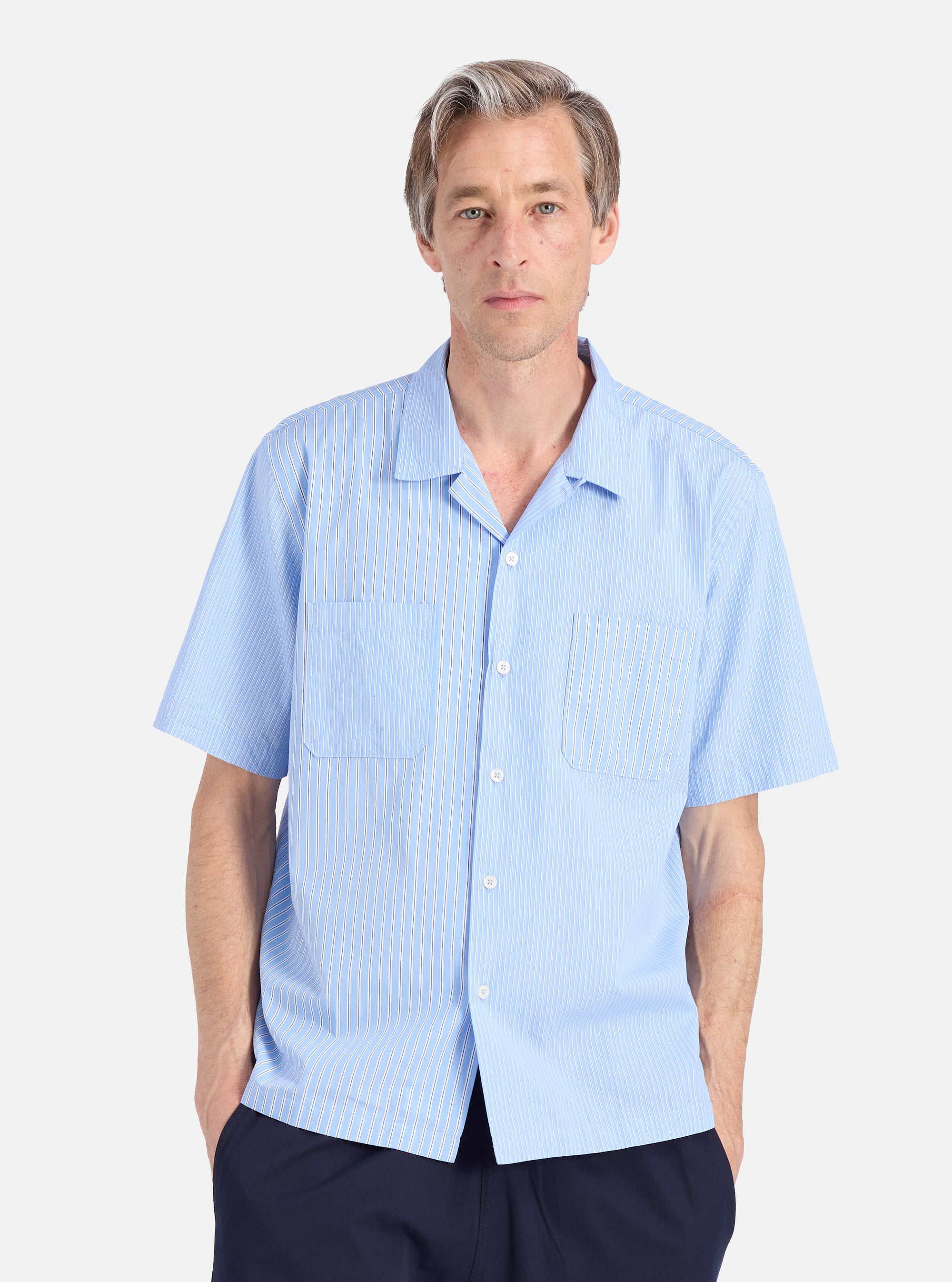 A man models the Blue Vila Stripes Sal Road Shirt. The light blue shirt has white stripes and short sleeves. Shot against a white backdrop.