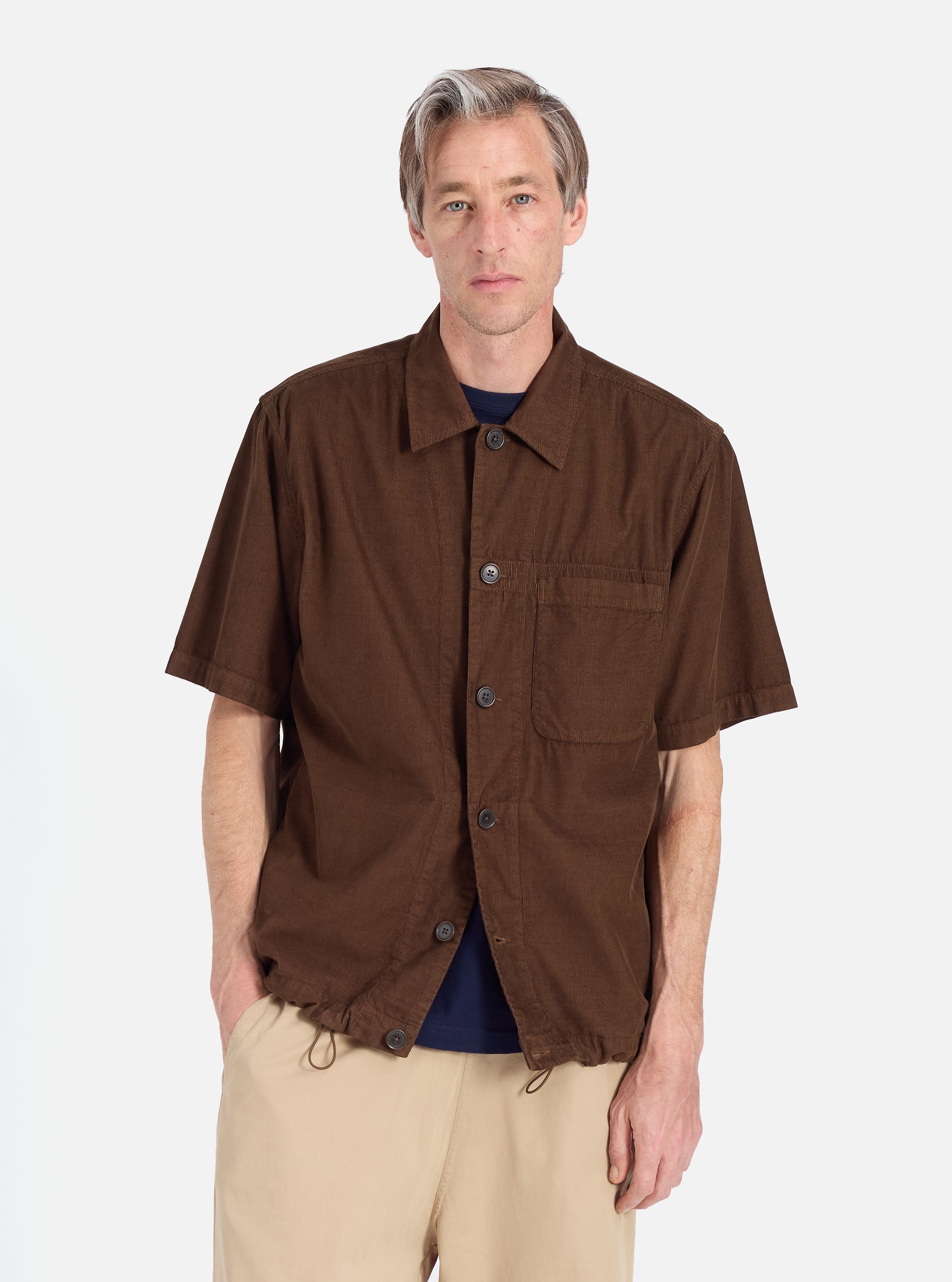 A model on a white background wears the Khaki Summer Cord Tech Overshirt, a brown corduroy short-sleeve shirt, paired with khaki pants. The overshirt has a relaxed fit.