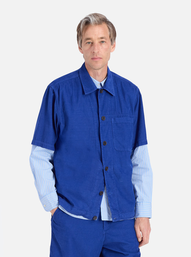 A man models the Work Blue Summer Cord Tech Overshirt against a white backdrop. The blue corduroy overshirt is worn over a striped shirt. The fit is relaxed and casual.