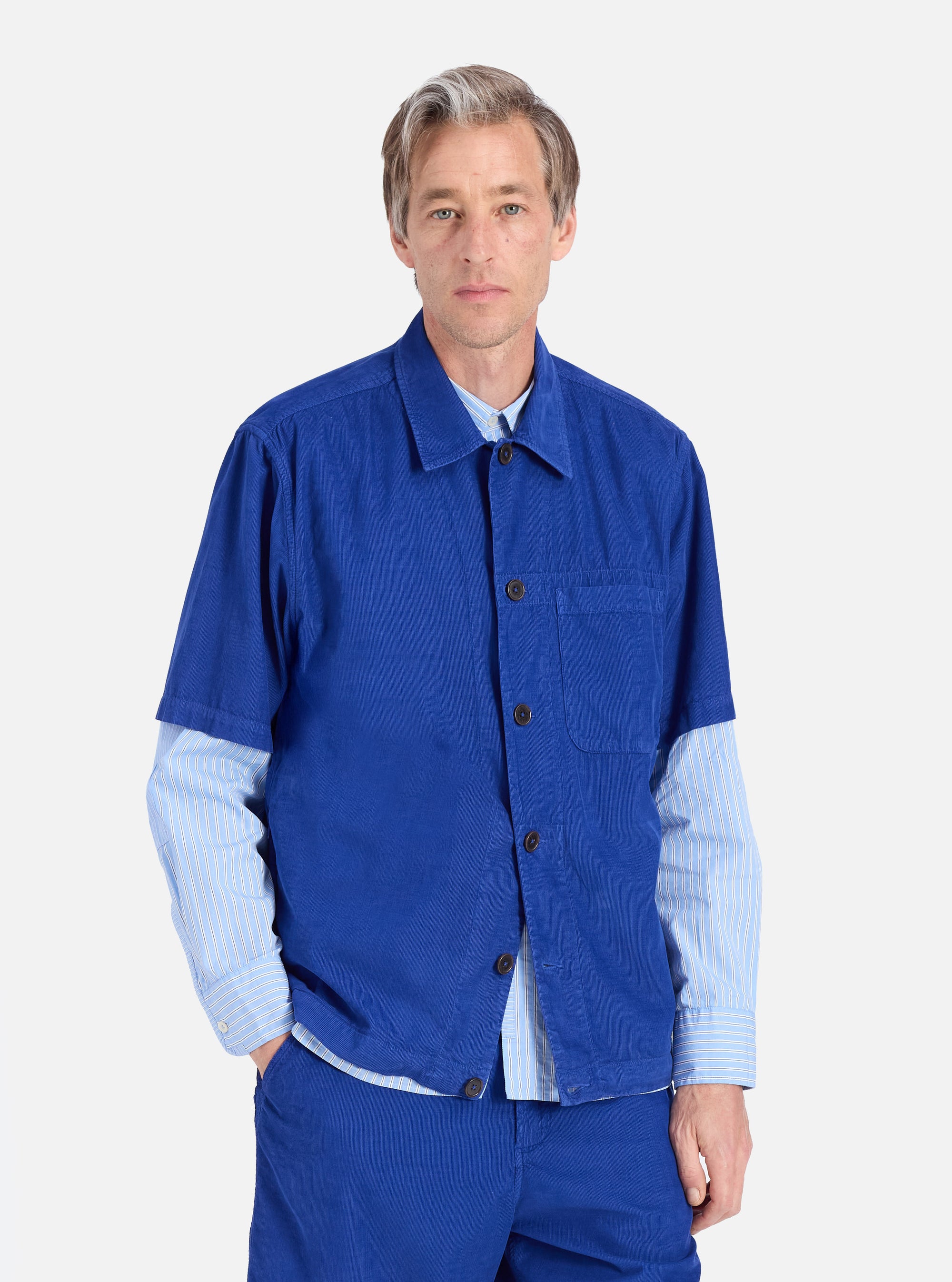 A man models the Work Blue Summer Cord Tech Overshirt against a white backdrop. The blue corduroy overshirt is worn over a striped shirt. The fit is relaxed and casual.