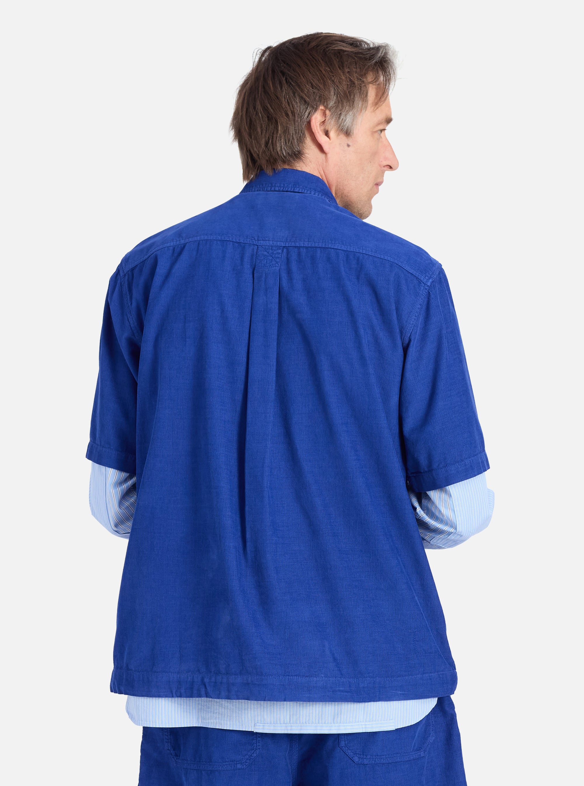 Back view of a man in the Work Blue Summer Cord Tech Overshirt. The corduroy overshirt is layered over a striped shirt. Shot against a white backdrop.