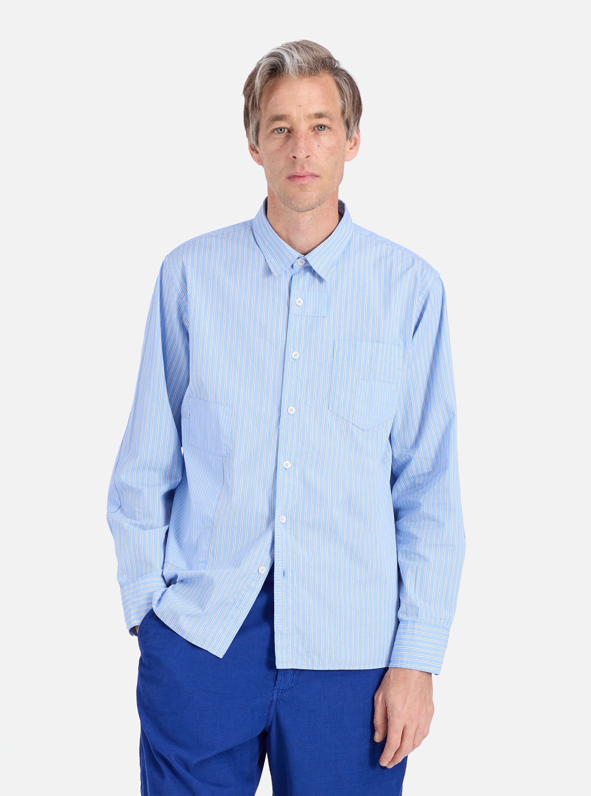 A man models the Blue Vila Stripes Patched Shirt against a white background. The shirt is light blue with white stripes. He wears blue pants. 