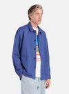 A man poses in a Blue Linen Cotton Shirting Travail Overshirt against a white background. The overshirt is buttoned over a graphic tee and paired with light wash pants.