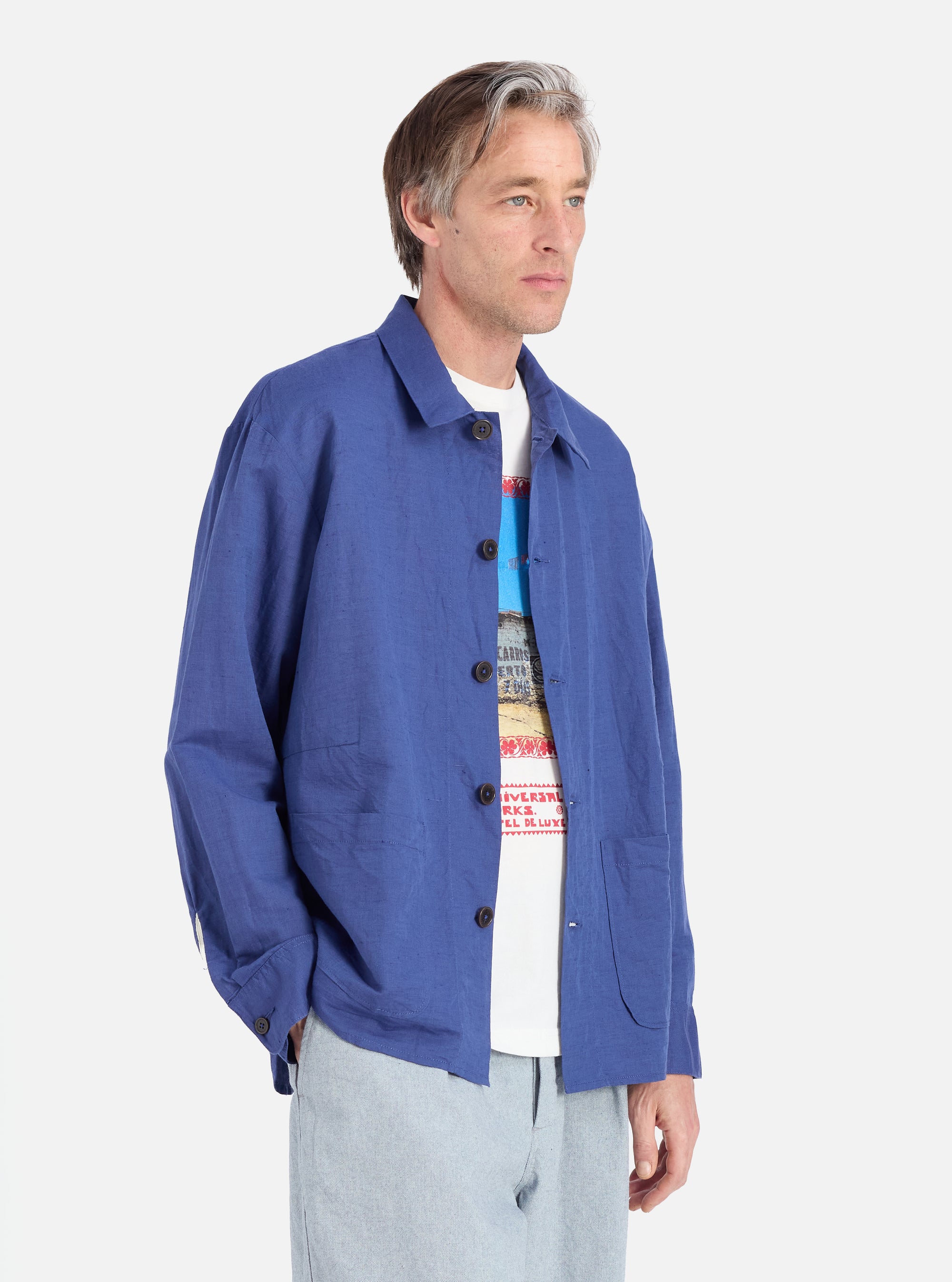 A man poses in a Blue Linen Cotton Shirting Travail Overshirt against a white background. The overshirt is buttoned over a graphic tee and paired with light wash pants.