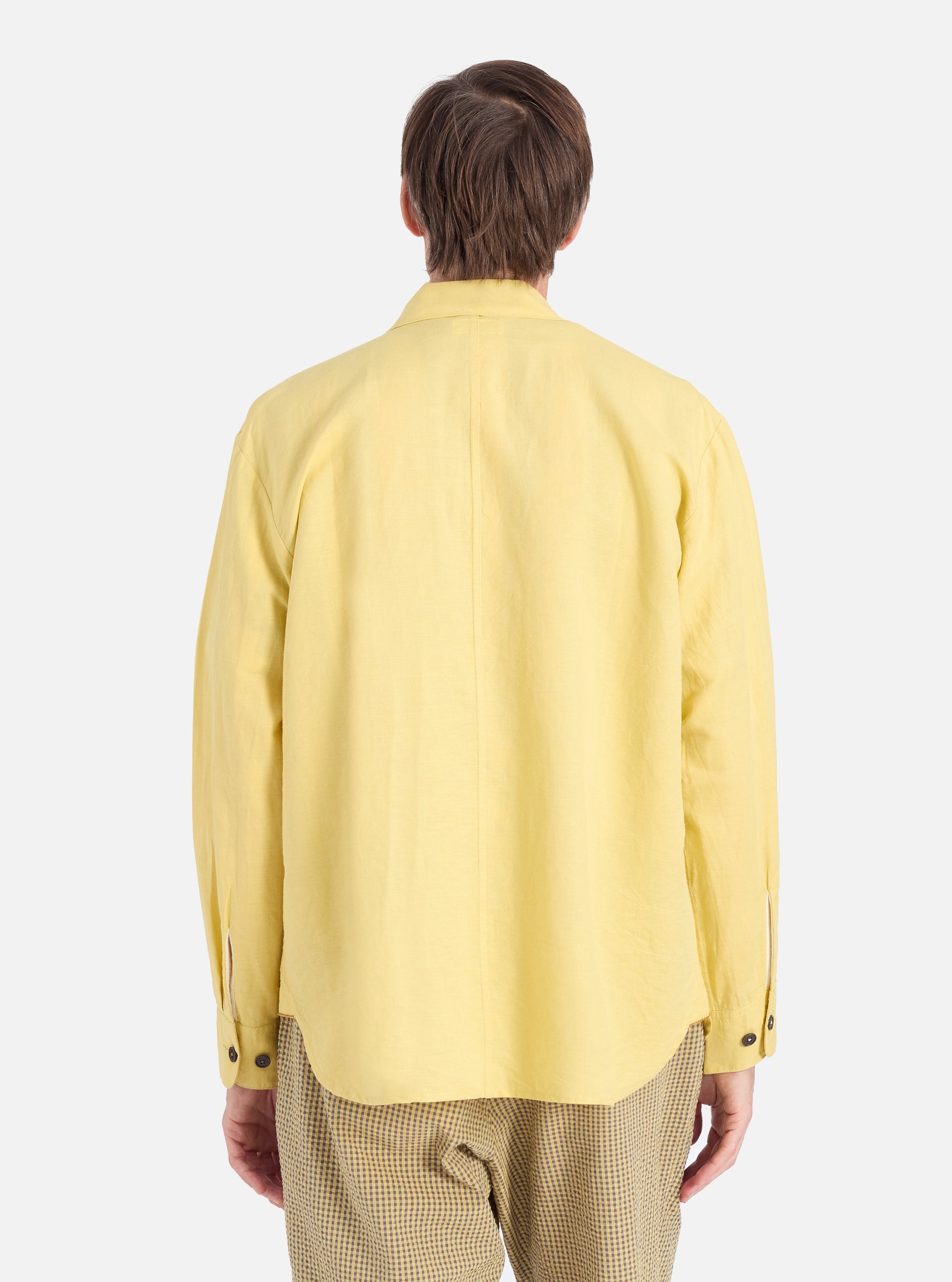 Back view of a model wearing a lemon-yellow linen cotton shirting Travail Overshirt. The overshirt is on a white background. The model is also wearing checkered pants.