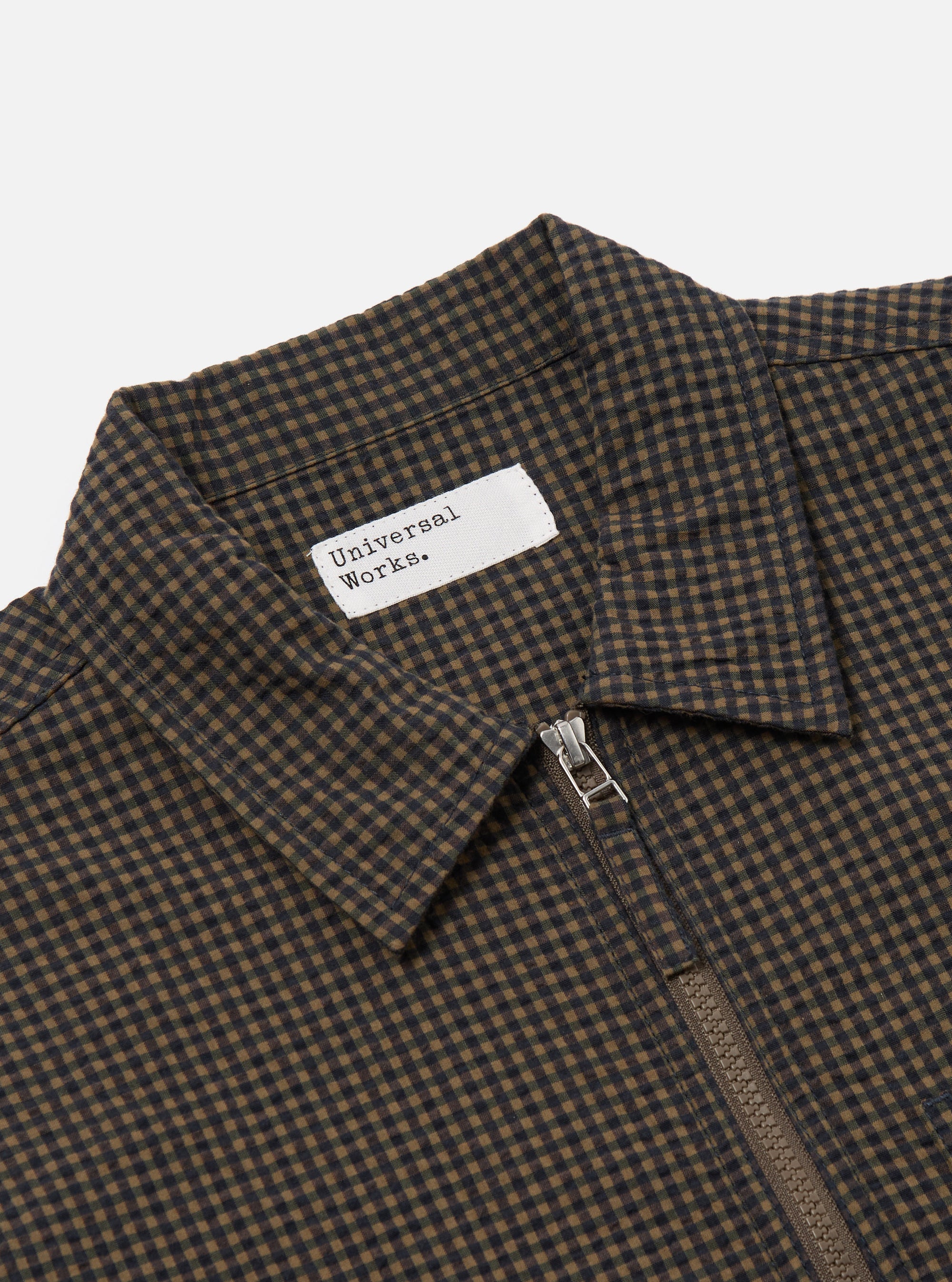 Close-up of the Navy Japanese Seersucker Zip Uniform Jacket. The jacket features a navy and brown checkered pattern, a silver zipper, and a white label. The background is a plain white.
