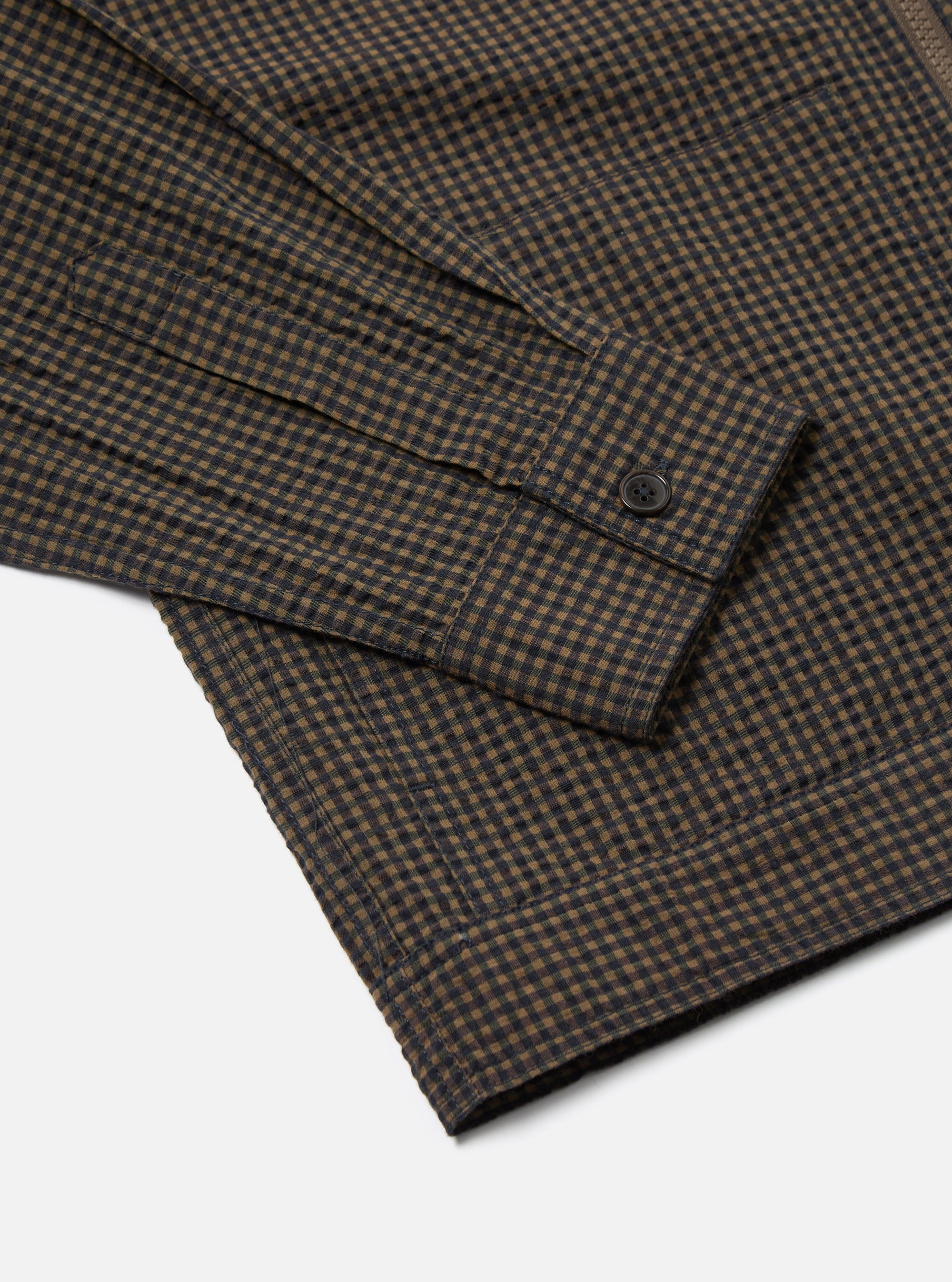 Close-up of the Navy Japanese Seersucker Zip Uniform Jacket showing the sleeve and hem. The jacket has a brown and navy checkered pattern. The background is a plain white.