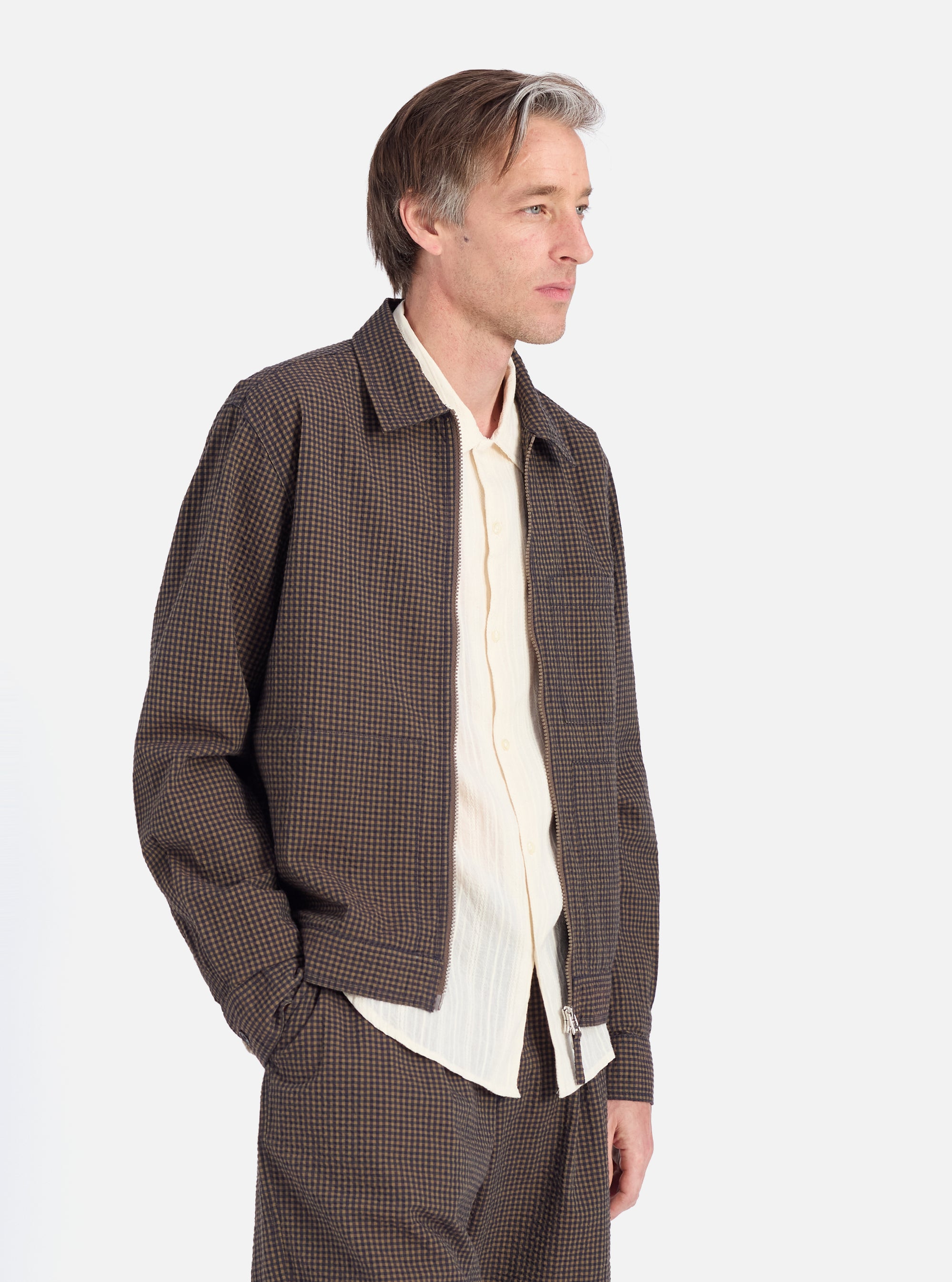 A man in profile on a white background wears the Navy Japanese Seersucker Zip Uniform Jacket. The jacket is brown and black check with a zip front. He wears a cream shirt underneath.
