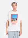 A model wears the Ecru/Red Single Jersey Pickup Print Tee against a white background. The tee is ecru with a red-framed print of a building. 