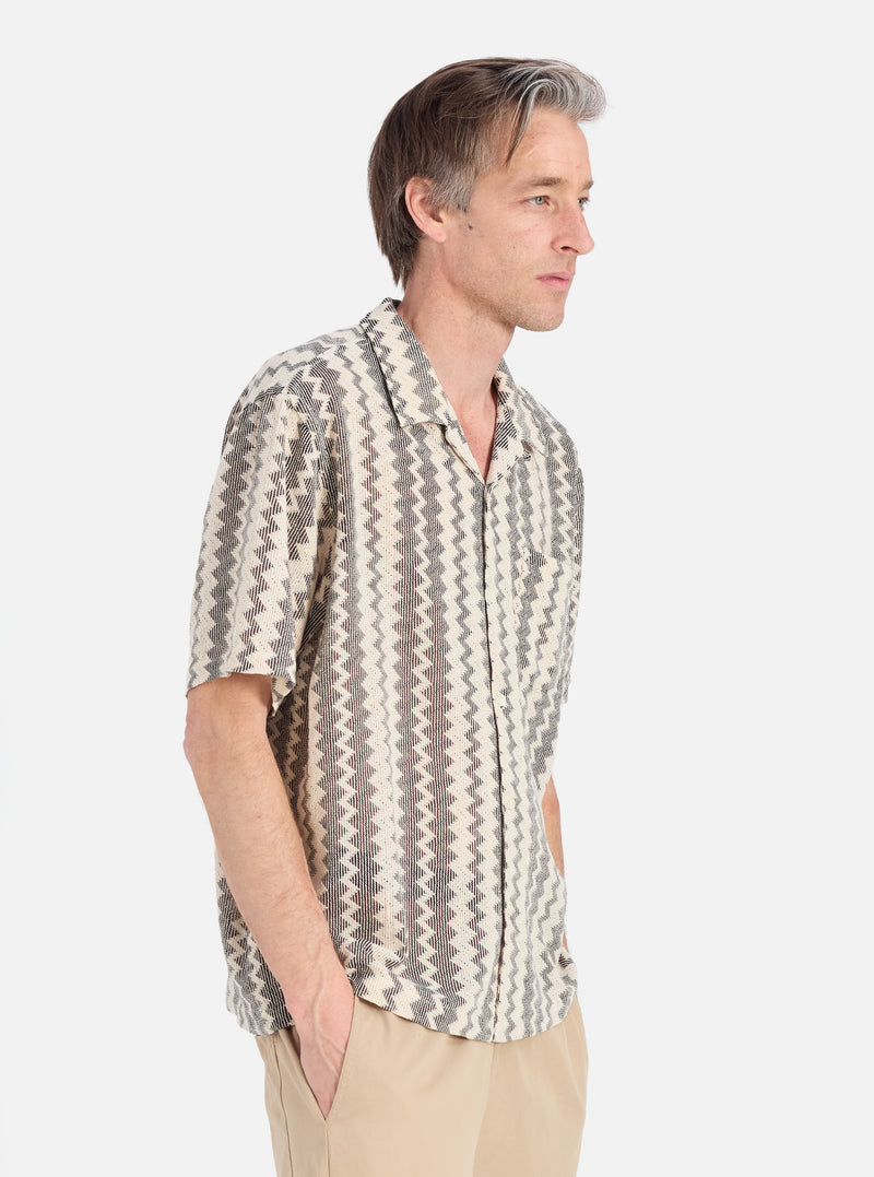 A man in profile on a white background wears the Charcoal Cotton Lattice Road Shirt. The shirt has a black and white geometric pattern. He wears khaki pants.