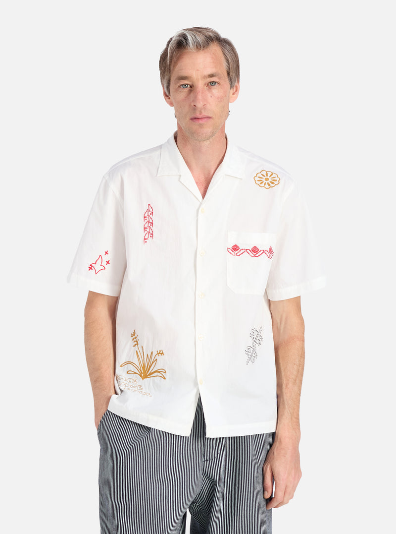 A model poses in the Ecru Embroidered Poplin Minari Shirt, a white poplin shirt with colorful embroidery. The background is a clean white.