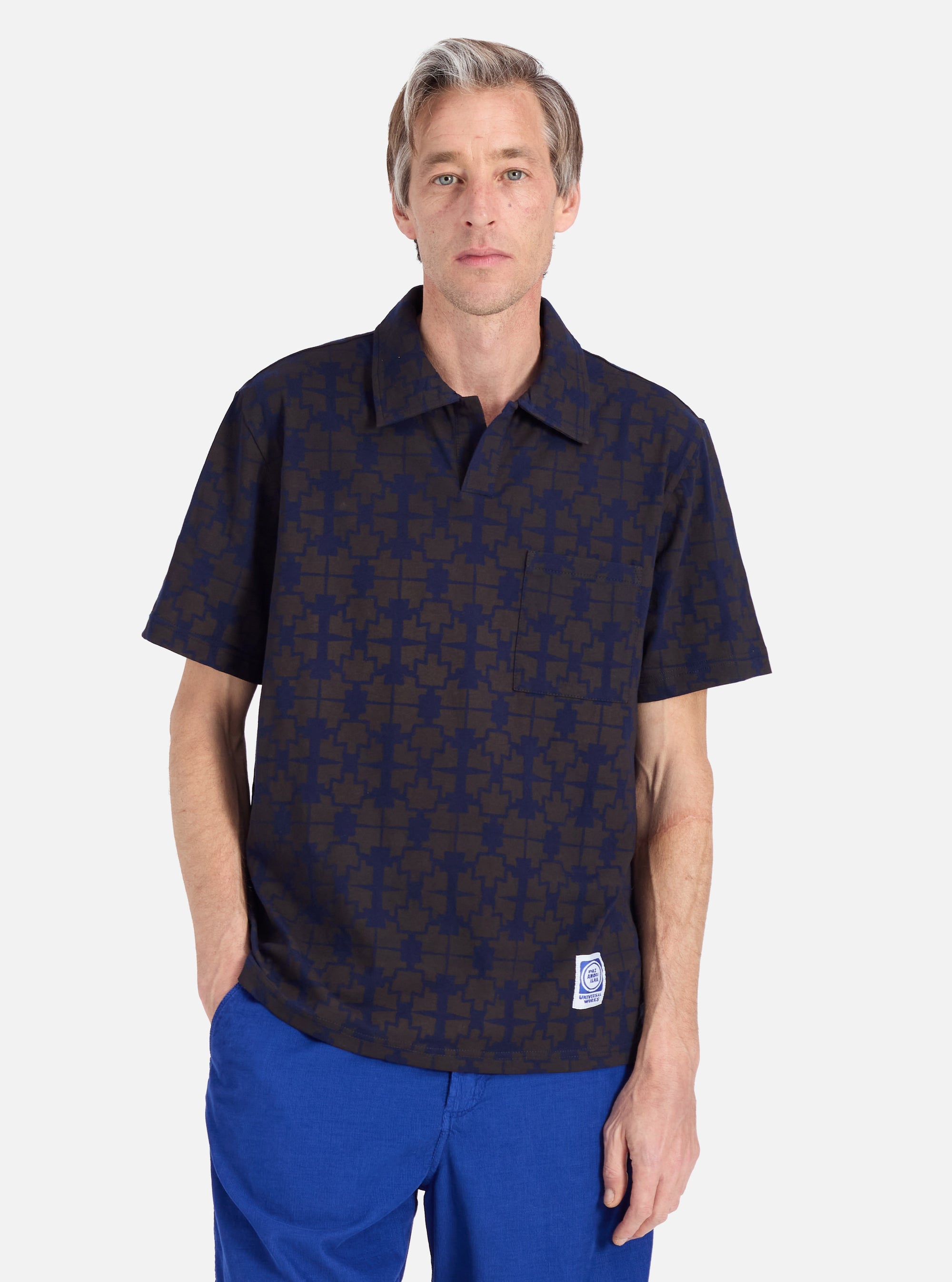 A model poses in front of a white background wearing the Navy/Brown Mitla Print Jersey Newlyn Polo. The polo has a navy and brown geometric print and short sleeves.