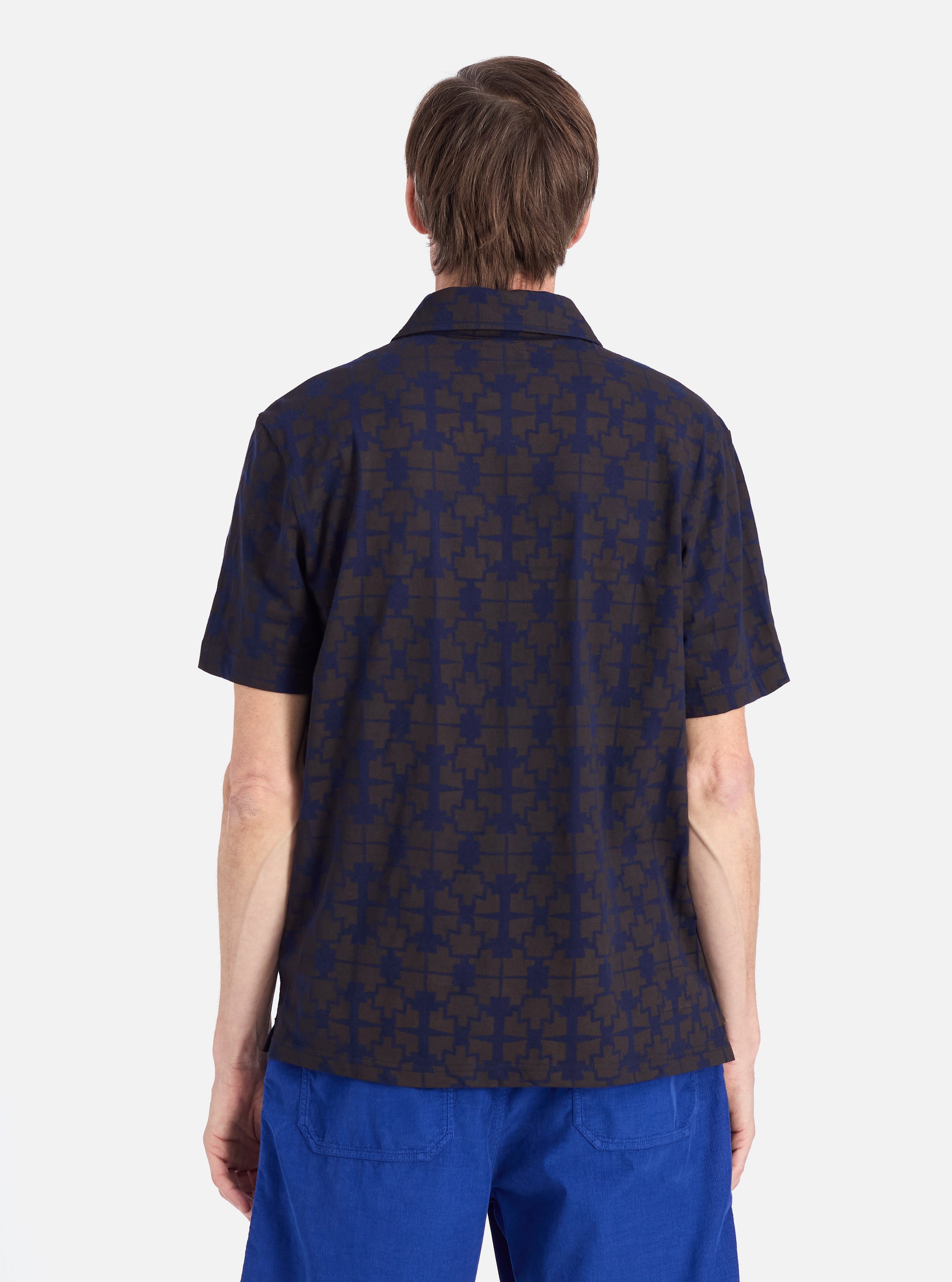 Back view of a model wearing a Navy/Brown Mitla Print Jersey Newlyn Polo on a white background. The polo features a navy and brown geometric print.