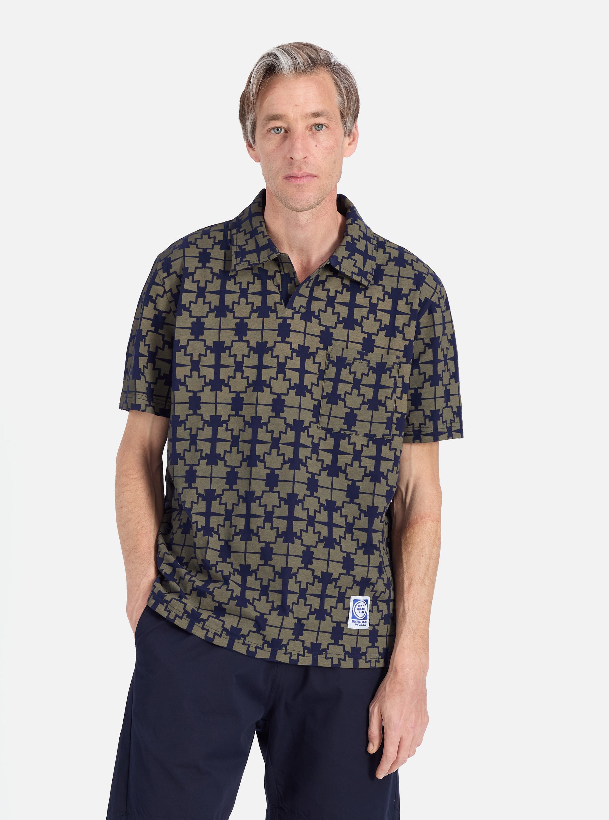 A man stands against a white backdrop, showcasing the Navy/Mud Mitla Print Jersey Newlyn Polo. The polo features a unique print and is paired with navy shorts.