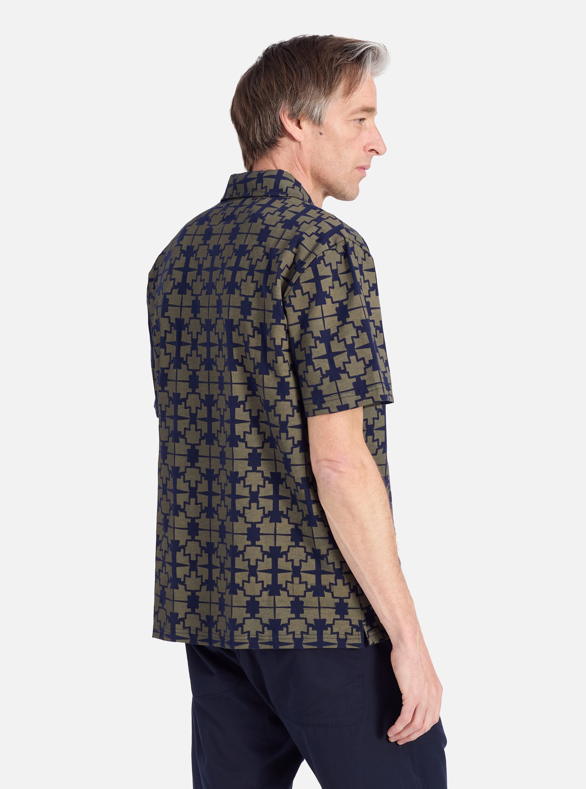 A man models the Navy/Mud Mitla Print Jersey Newlyn Polo against a white backdrop. The polo features a unique navy and mud-colored geometric print.