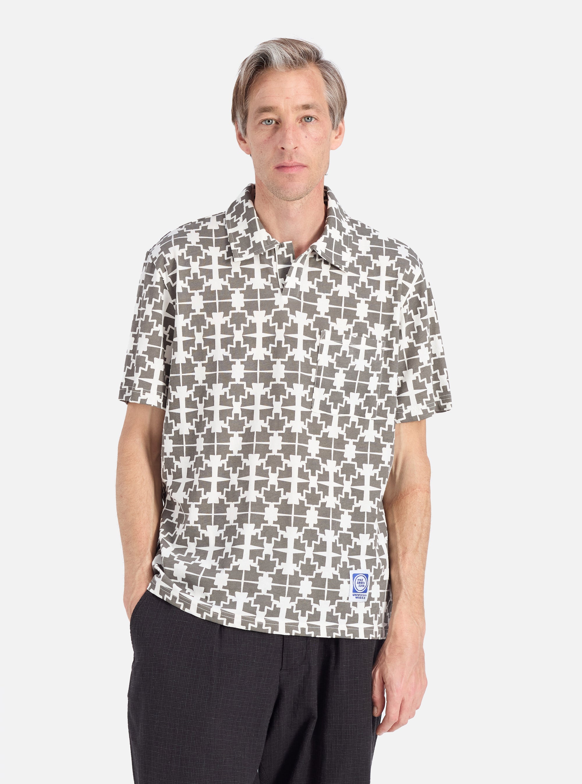 A model poses in the Ecru/Mud Mitla Print Jersey Newlyn Polo. The polo is ecru with a mud-colored geometric print. The background is a clean, light grey.
