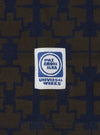 Close-up of the Navy/Brown Mitla Print Jersey Newlyn Polo fabric, featuring a geometric pattern. A Universal Works label is stitched on. 