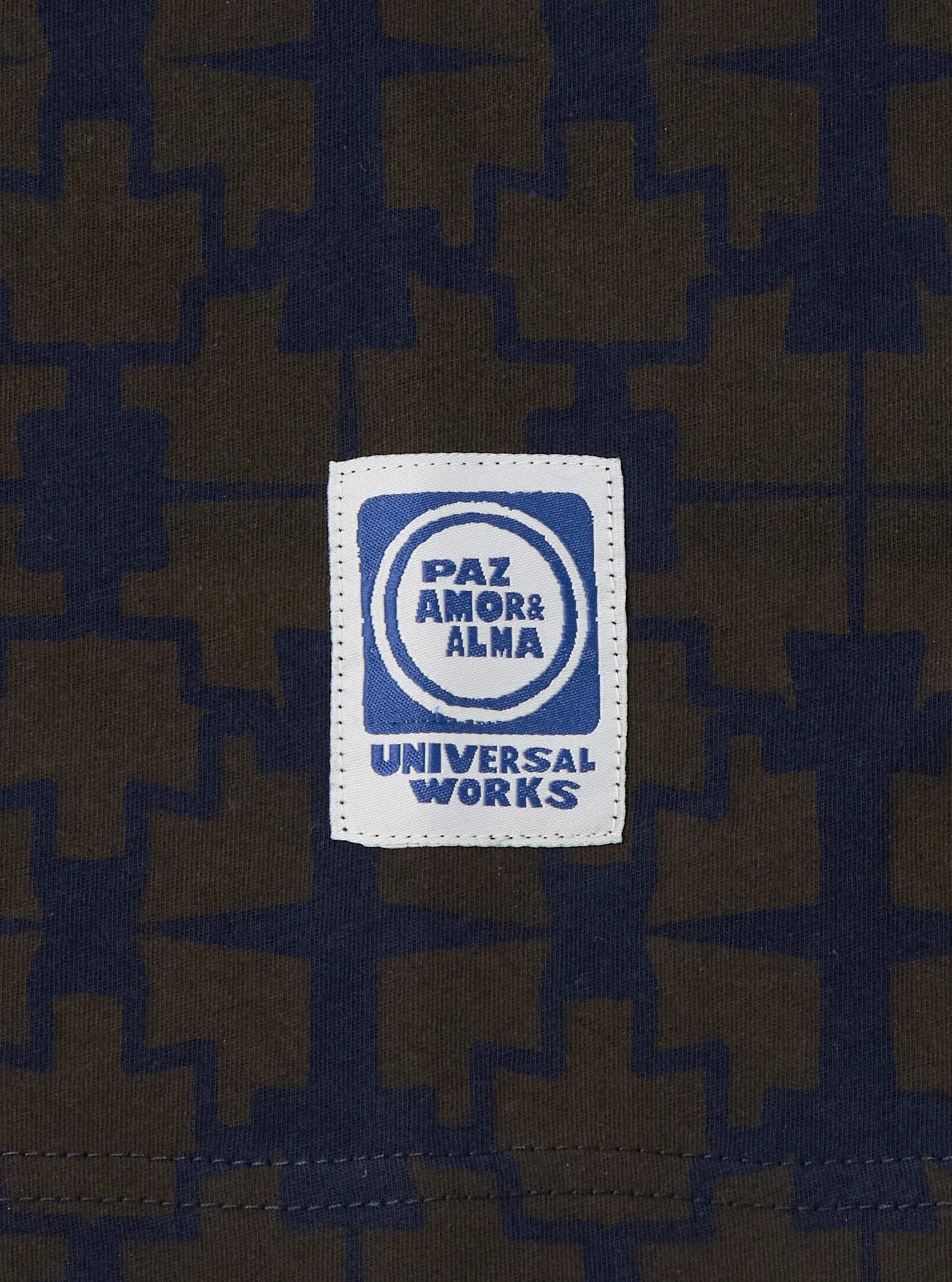 Close-up of the Navy/Brown Mitla Print Jersey Newlyn Polo fabric, featuring a geometric pattern. A Universal Works label is stitched on. 