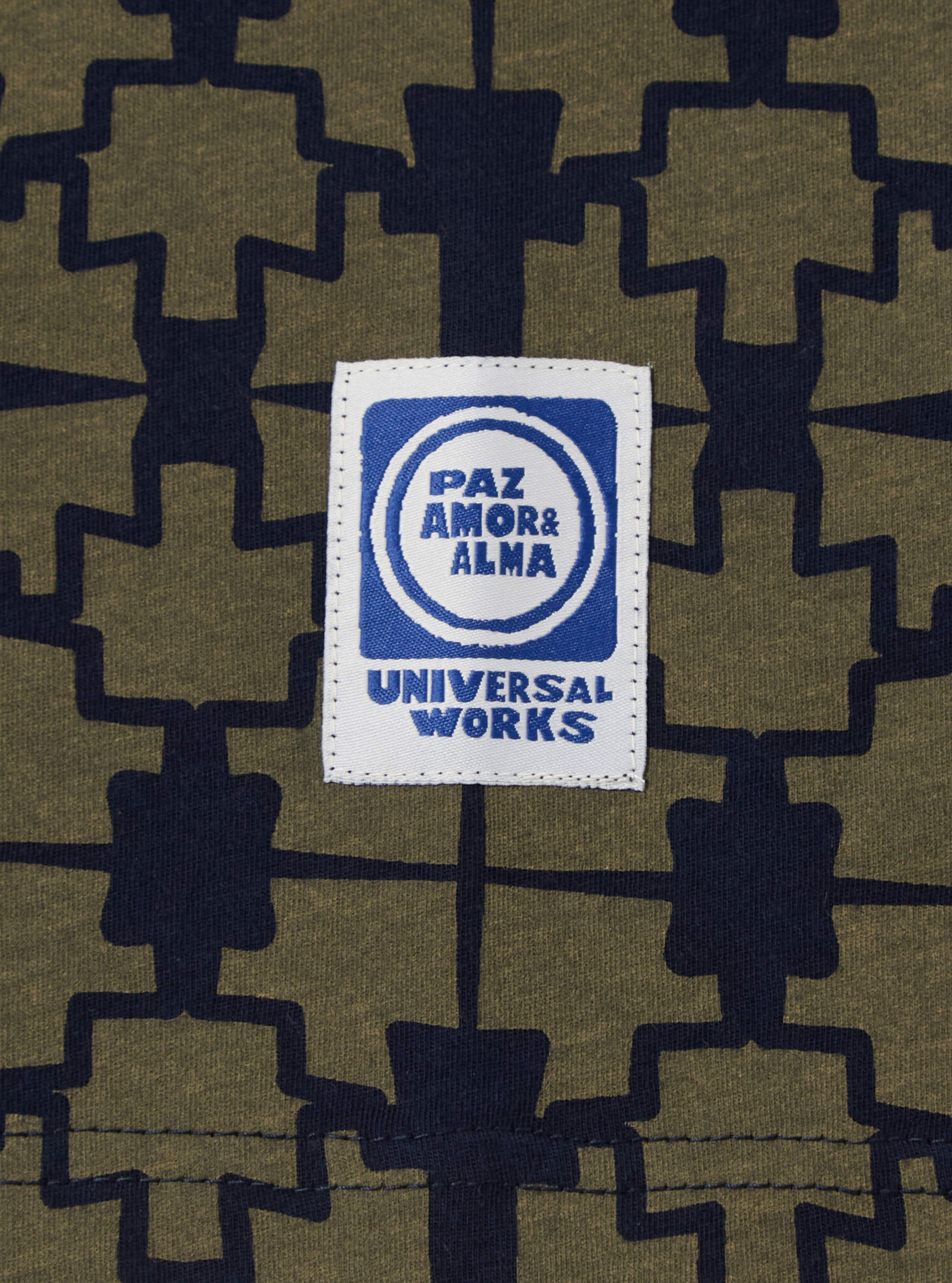 Close-up of the Navy/Mud Mitla Print Jersey Newlyn Polo fabric, showcasing the intricate pattern and the 'Universal Works' label. The jersey material is soft and textured.