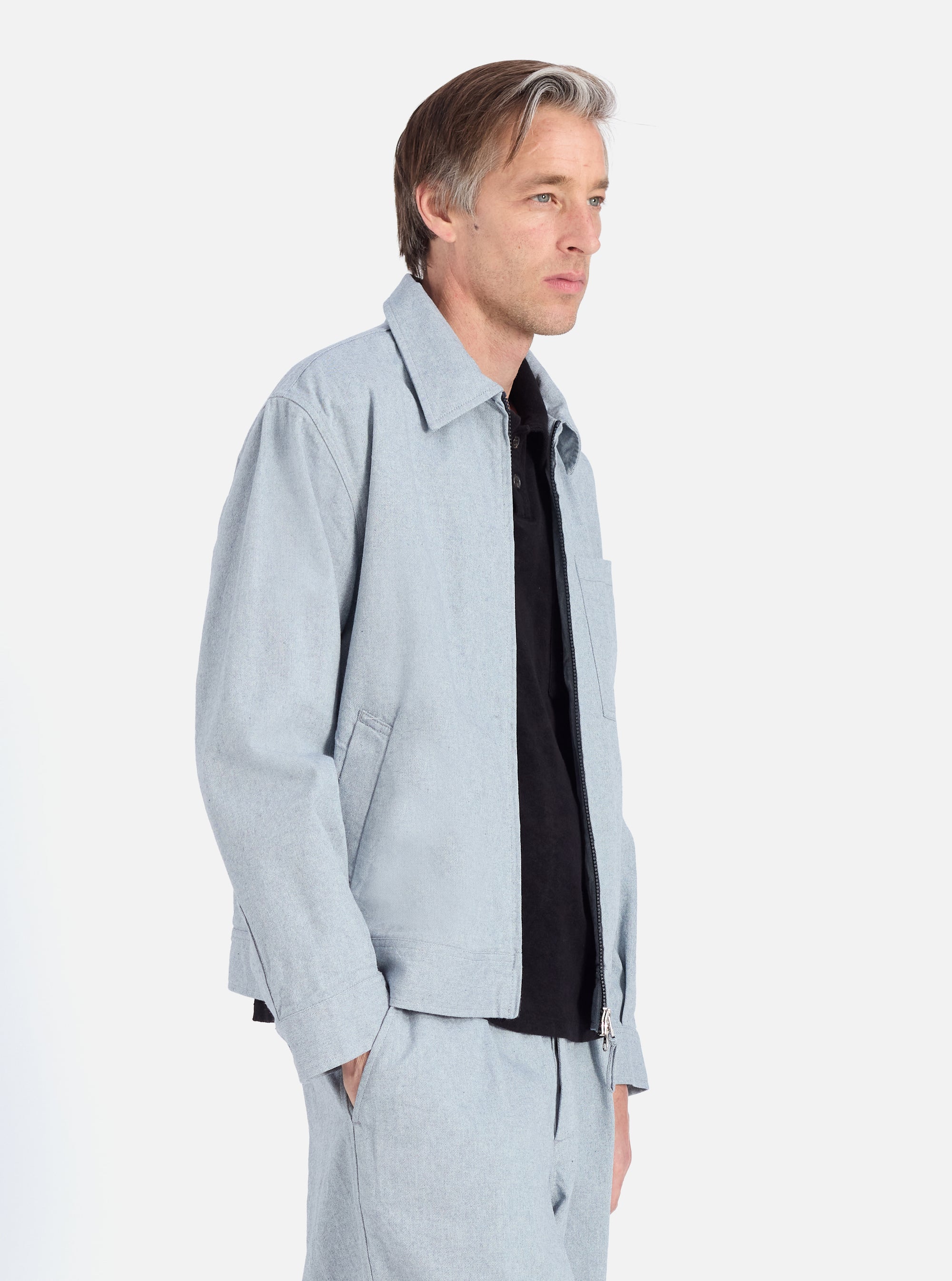 A man poses in the Indigo Reworked Jean Cloth Rose Bowl Jacket. The light blue jacket is denim-like. He wears a black shirt. Studio shot on white background.