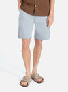 A model on a white background showcases the Indigo Reworked Jean Cloth Alva Short. The light blue shorts are paired with a brown shirt and sandals for a casual look.