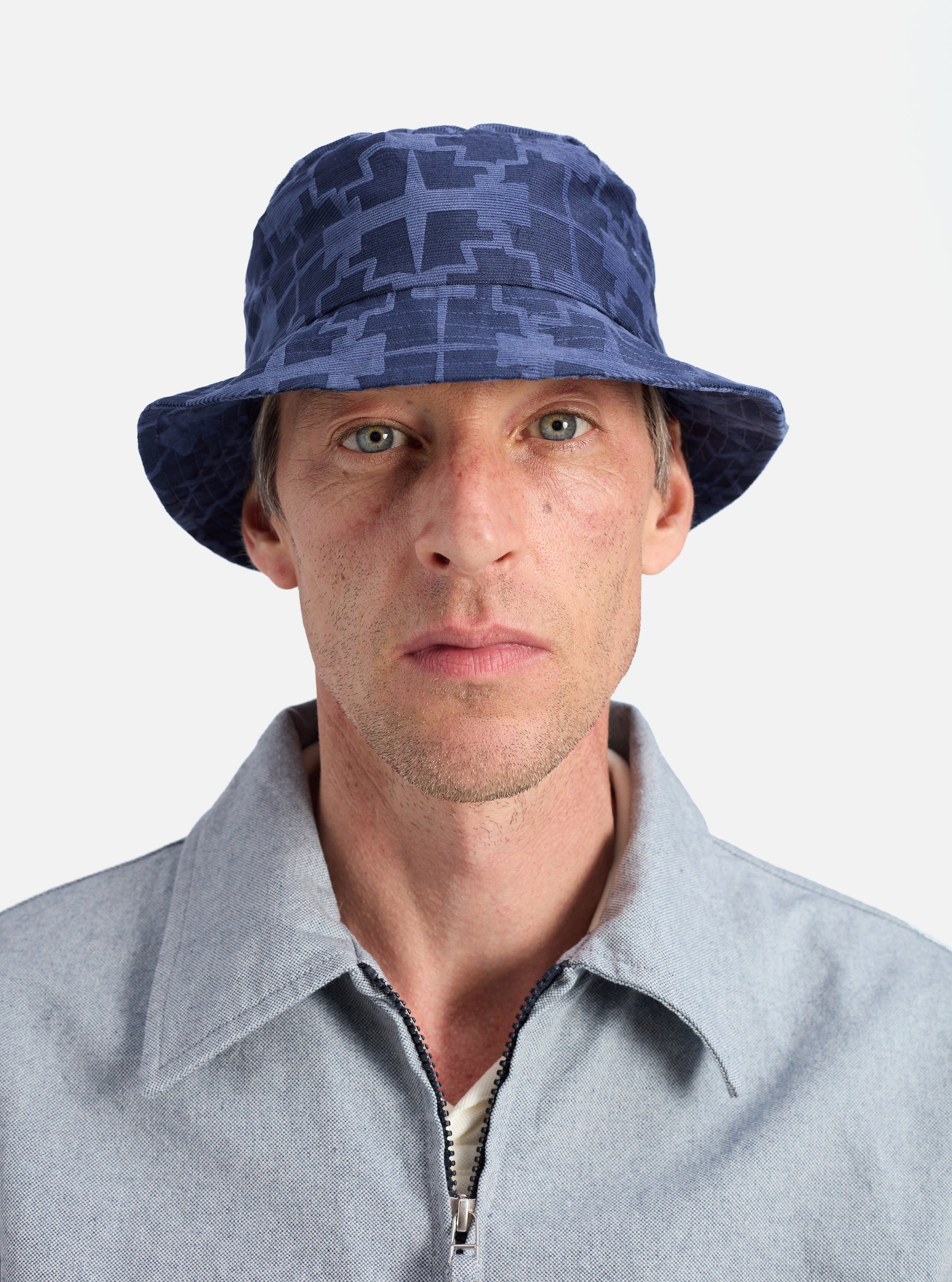 A model poses in front of a white background wearing a Navy Mitla Print Cord Beach Hat. The hat is blue corduroy with a geometric print.
