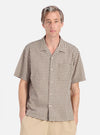 A man models the Olive Dobby Check Road Shirt against a white backdrop. The shirt features a brown and white check pattern with short sleeves and a relaxed fit.
