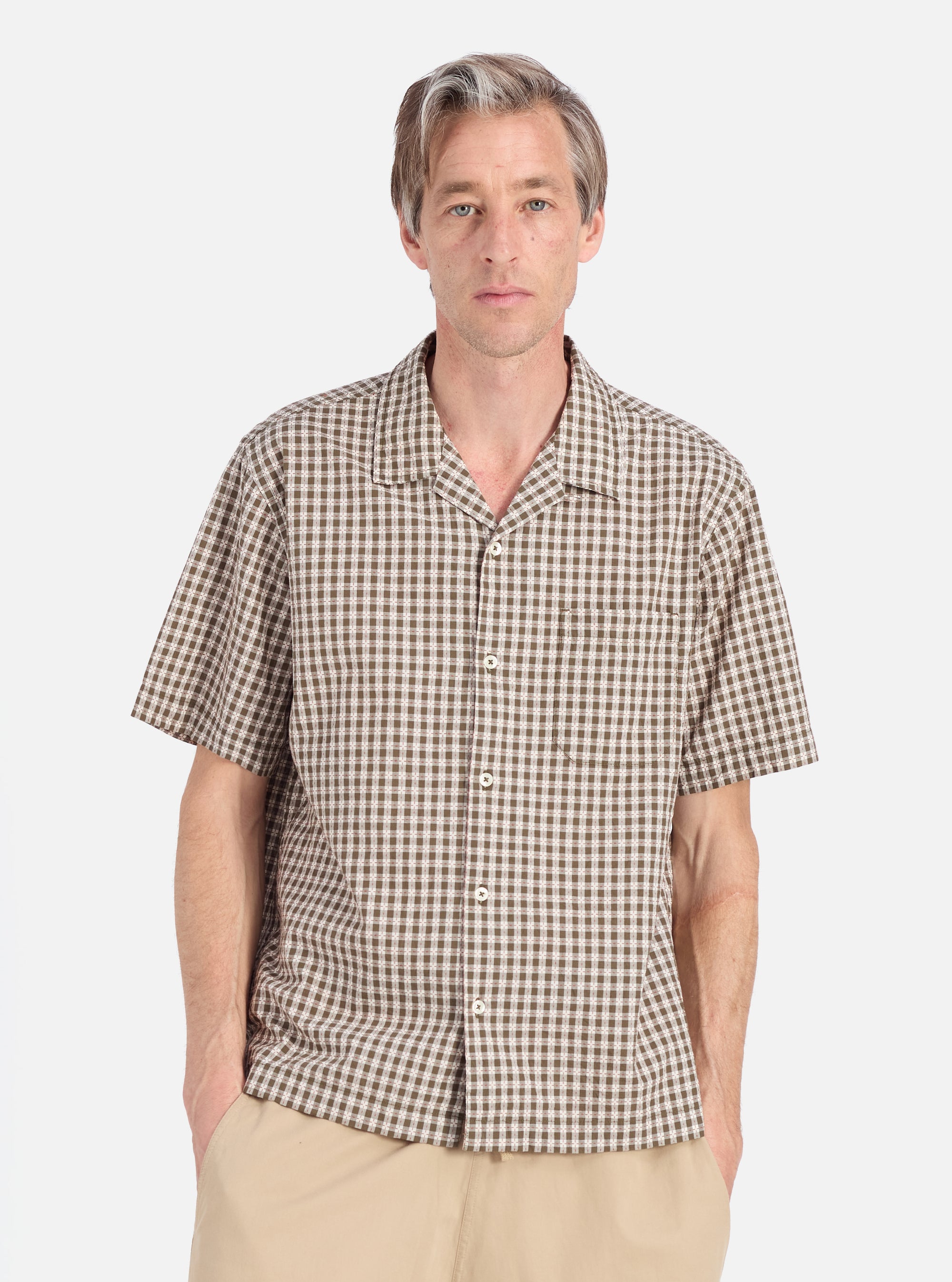A man models the Olive Dobby Check Road Shirt against a white backdrop. The shirt features a brown and white check pattern with short sleeves and a relaxed fit.