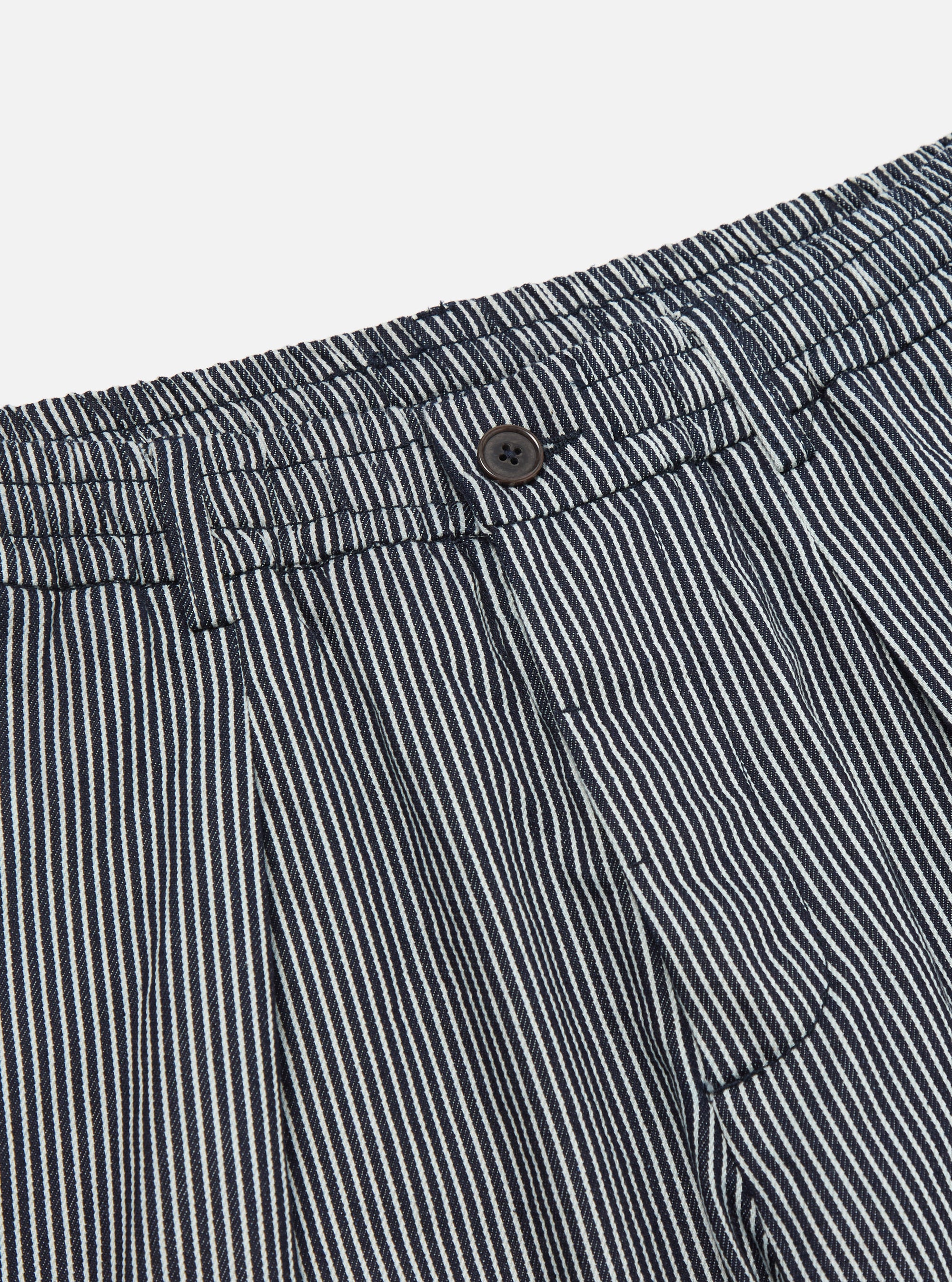 Close-up of the Indigo Hickory Stripe Denim Pleated Track Pant. Features include a button closure, pleated design, and denim material. Shot on a plain white background.
