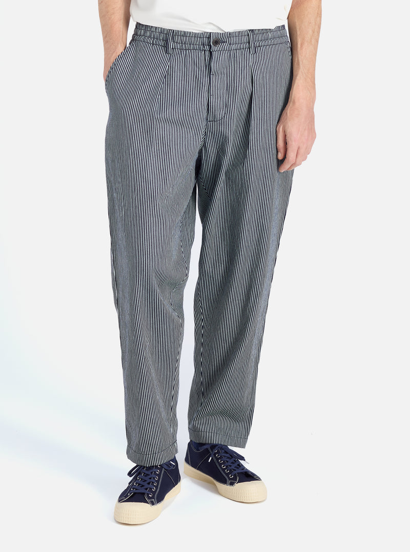 A man stands in front of a white background modeling the Indigo Hickory Stripe Denim Pleated Track Pant. The striped pants are paired with blue sneakers.