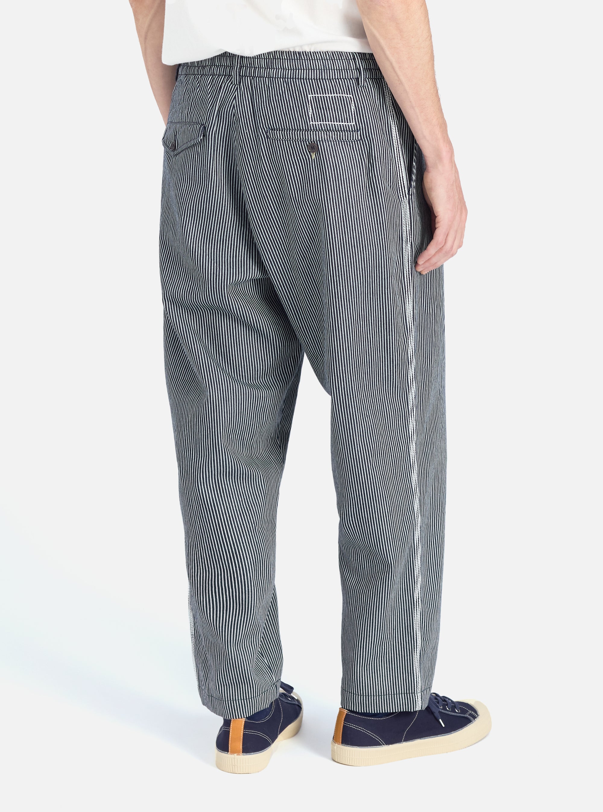 Back view of a model wearing Indigo Hickory Stripe Denim Pleated Track Pant. The denim pants have a relaxed fit. Shot against a white background.