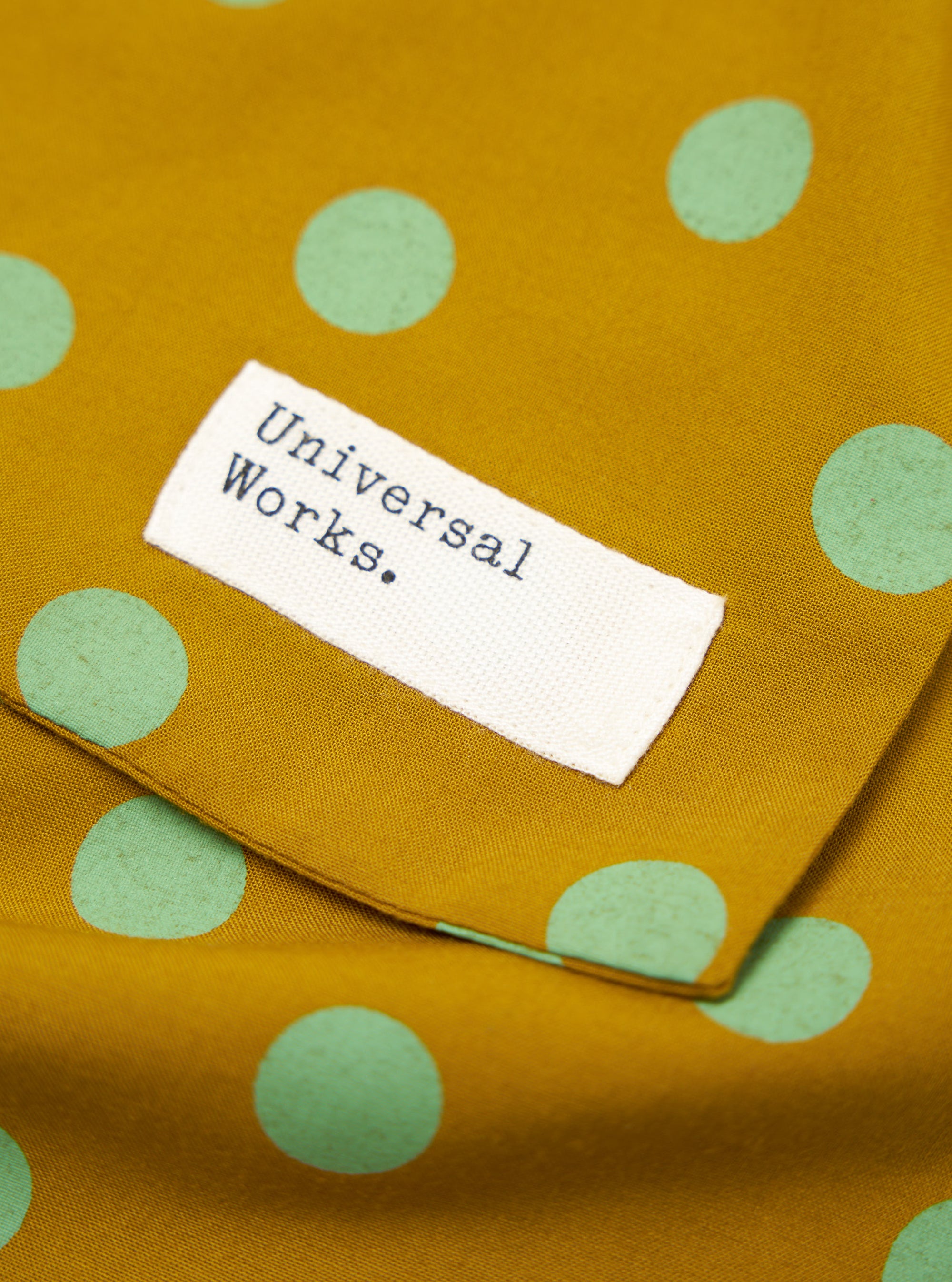 Close-up of a Mustard Bold Dot Print Rayon Evening Scarf. The scarf is mustard colored with light green polka dots. A white tag with "Universal Works." is sewn on.