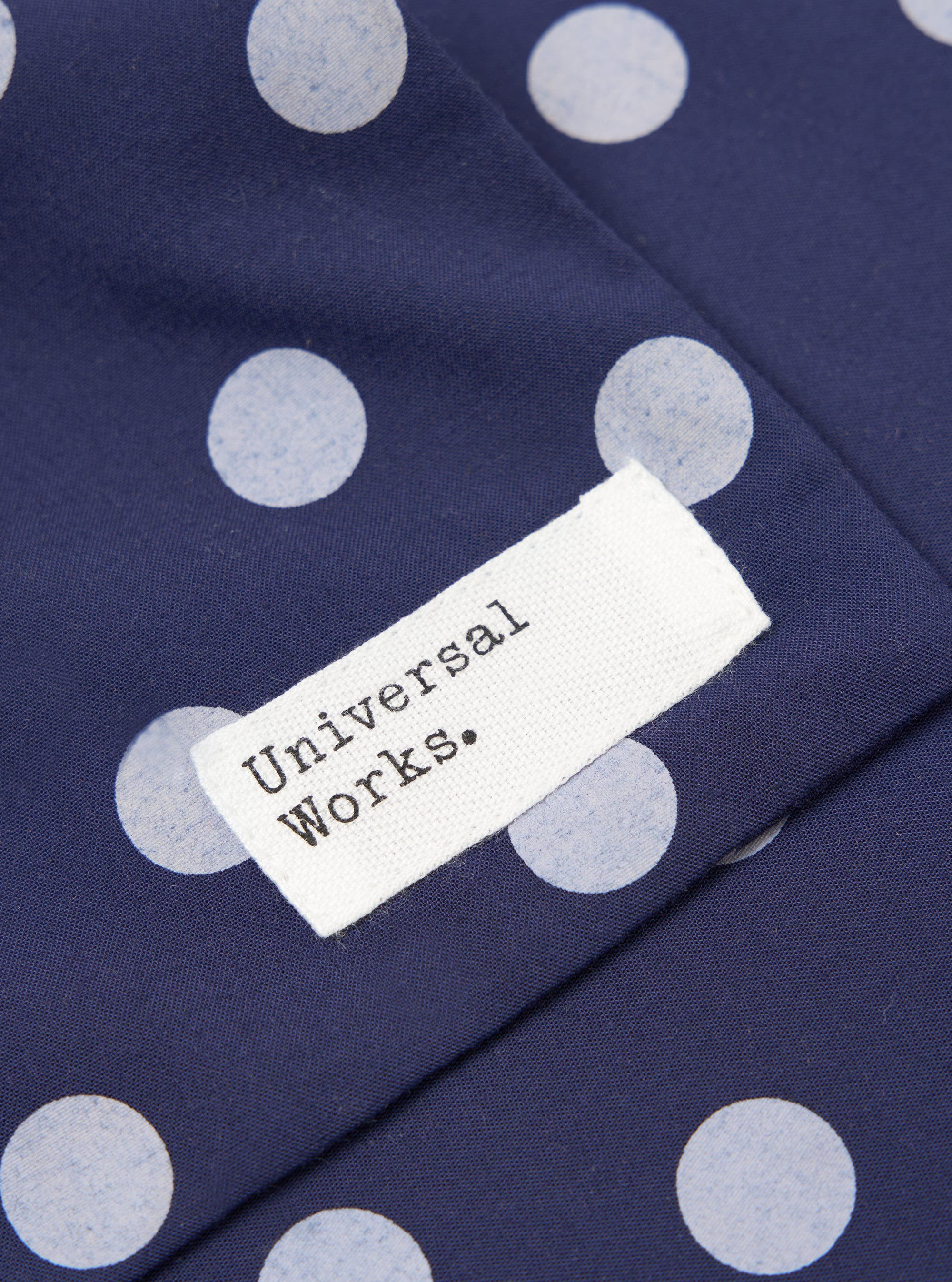Close-up of the Navy Bold Dot Print Rayon Evening Scarf featuring a navy fabric with white polka dots and a white tag with the brand name 'Universal Works.'