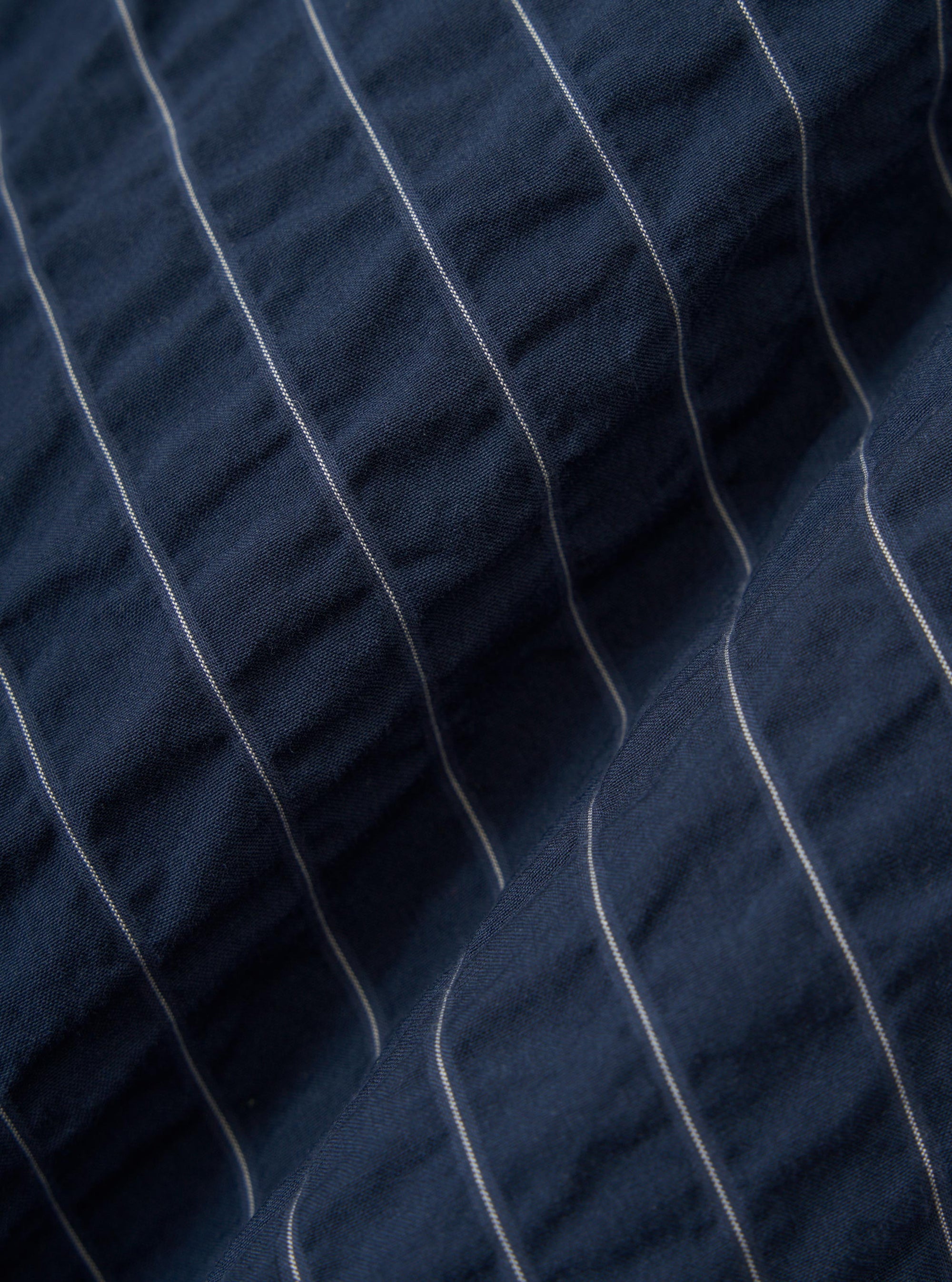 A close-up fabric swatch of the Navy Chester Stripe Two Button Jacket displays a detailed view of the navy blue cotton material with fine, vertical white stripes. The image highlights the texture and weave of the fabric.