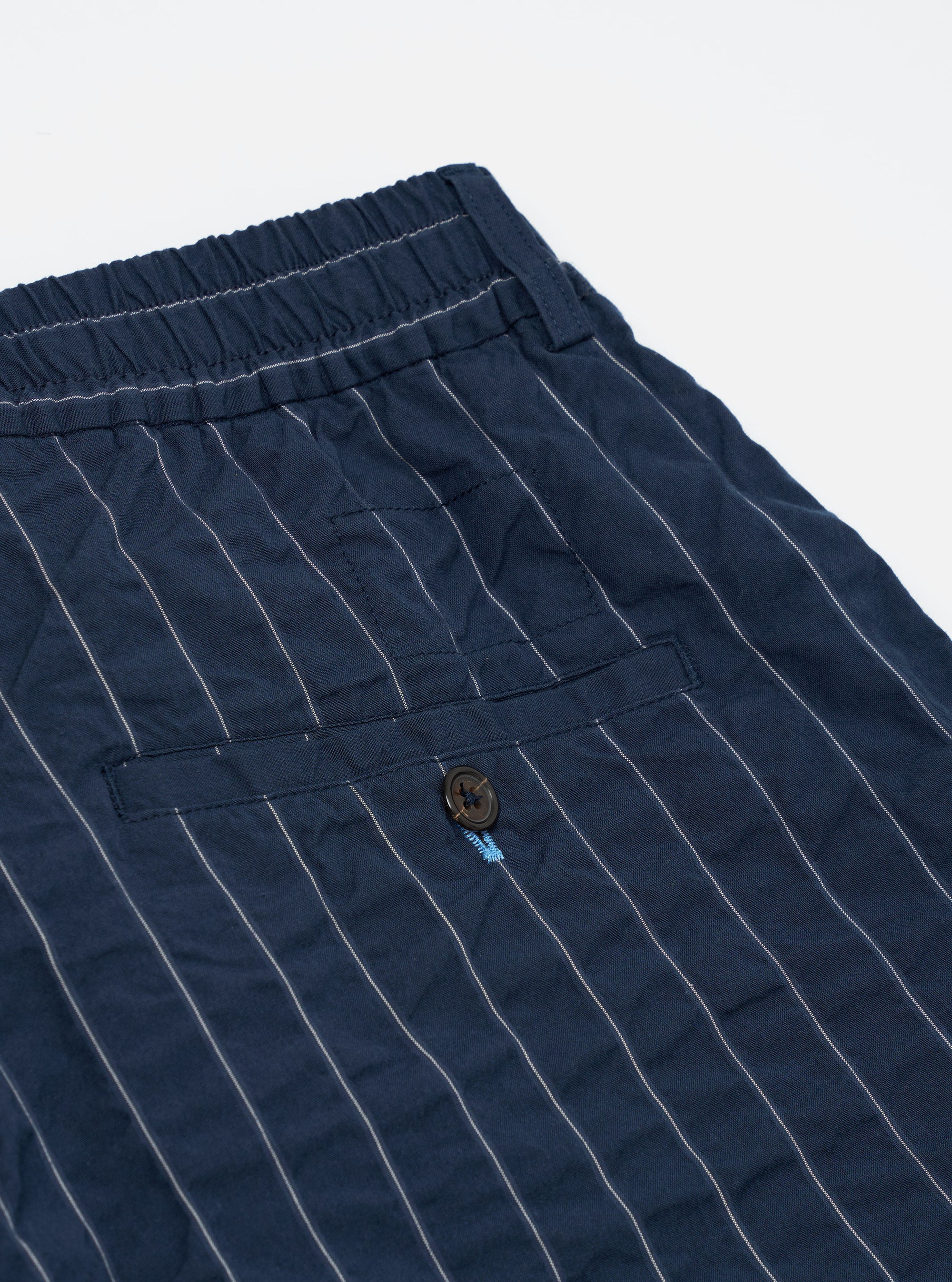 The Navy Chester Stripe Pleated Track Short is shown from a rear perspective, laid flat against a plain white background. The image highlights the two rear pockets, one of which is a buttoned jetted pocket, and the elasticated waistband. The fabric features a dark navy and white pinstriped seersucker texture.