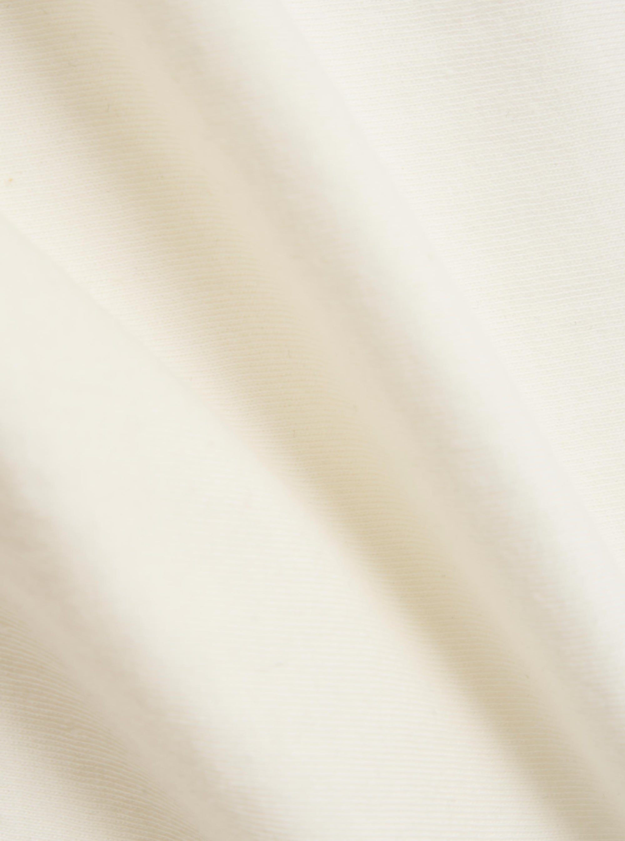 A close-up fabric swatch of the Ecru Single Jersey Multi UW Print Multi UW Print Tee is shown. The image features the ecru-colored cotton jersey material with a portion of a multi-colored logo print visible. The fabric has a fine, knitted texture.