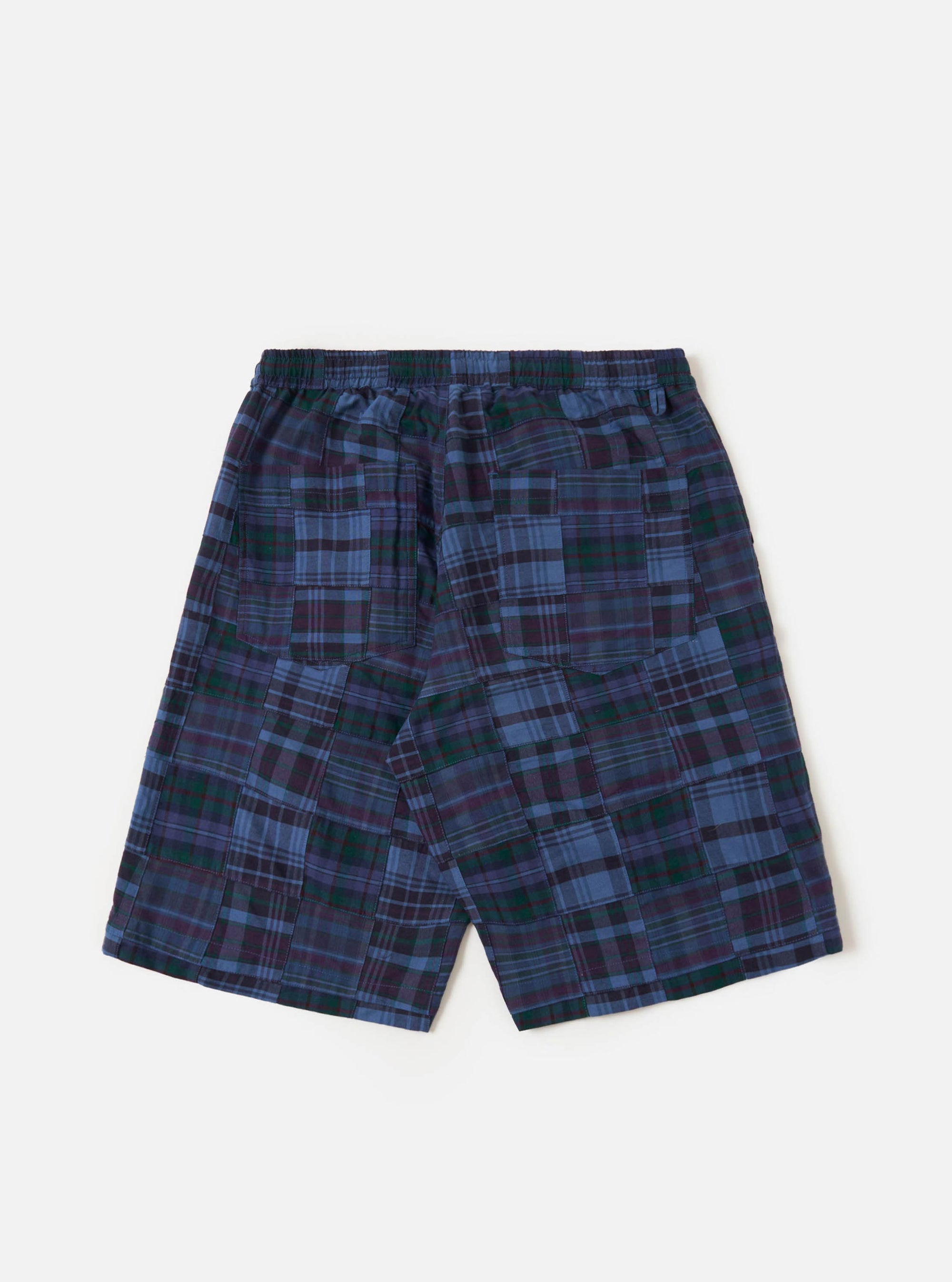 A pair of navy madras patchwork vacation shorts is shown from a rear angle, laid flat against a plain white background. The image highlights the patchwork construction of various blue and navy plaid patterns, the elasticated waistband with a drawstring, and a single rear patch pocket on the right side.