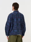 A model is wearing the Navy Madras Patchwork Duty Jacket against a plain white background. The image, taken from a side-rear perspective, highlights the jacket's oversized fit and features a vibrant blue and navy plaid patchwork pattern. The model is shown from the waist up, showcasing the curved hem and darted sleeves.