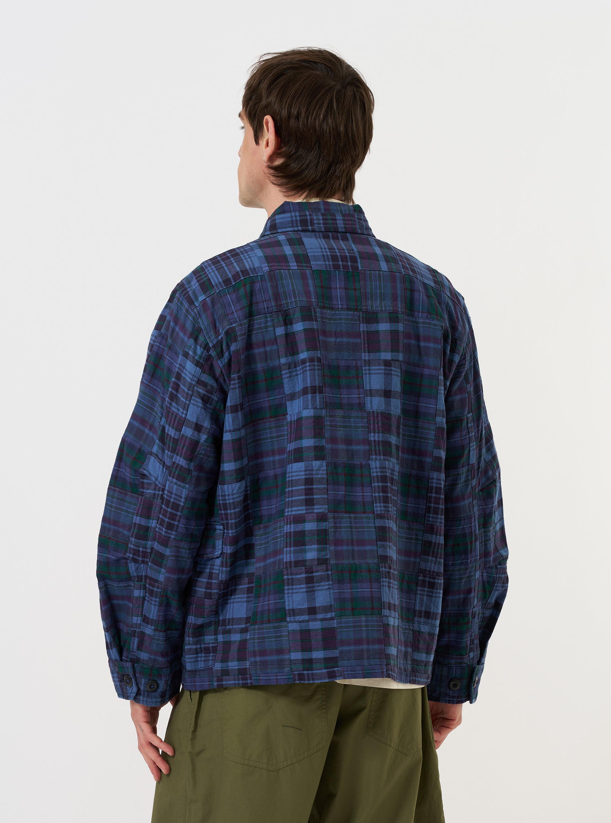 A model is wearing the Navy Madras Patchwork Duty Jacket against a plain white background. The image, taken from a side-rear perspective, highlights the jacket's oversized fit and features a vibrant blue and navy plaid patchwork pattern. The model is shown from the waist up, showcasing the curved hem and darted sleeves.