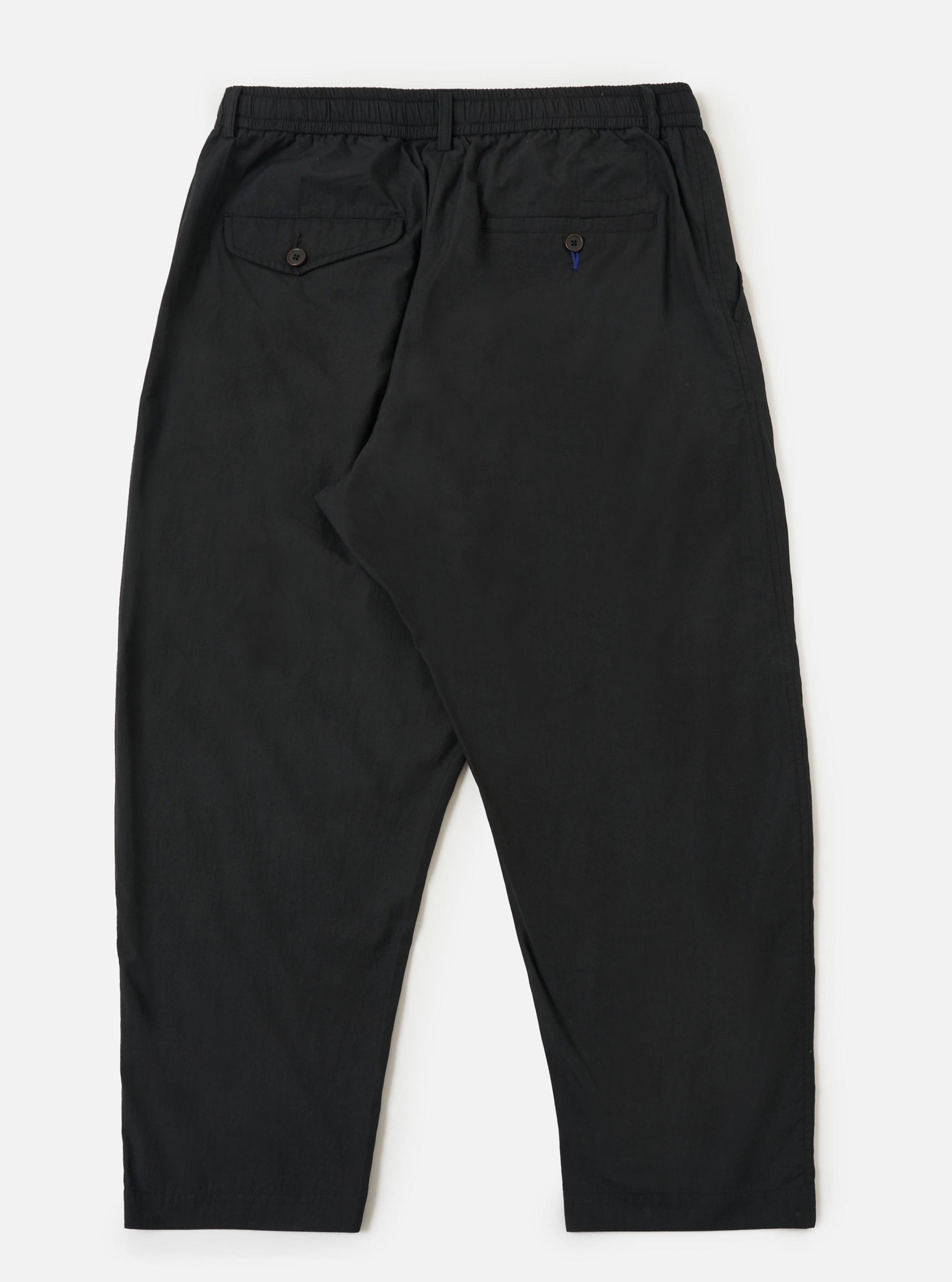 A pair of Black Broadcloth Oxford Pants is shown from the back, laid flat against a plain white background. The image highlights the two rear pockets—one buttoned flap pocket and one buttoned jetted pocket—as well as the elasticated waistband and the wide-leg cut of the trousers.