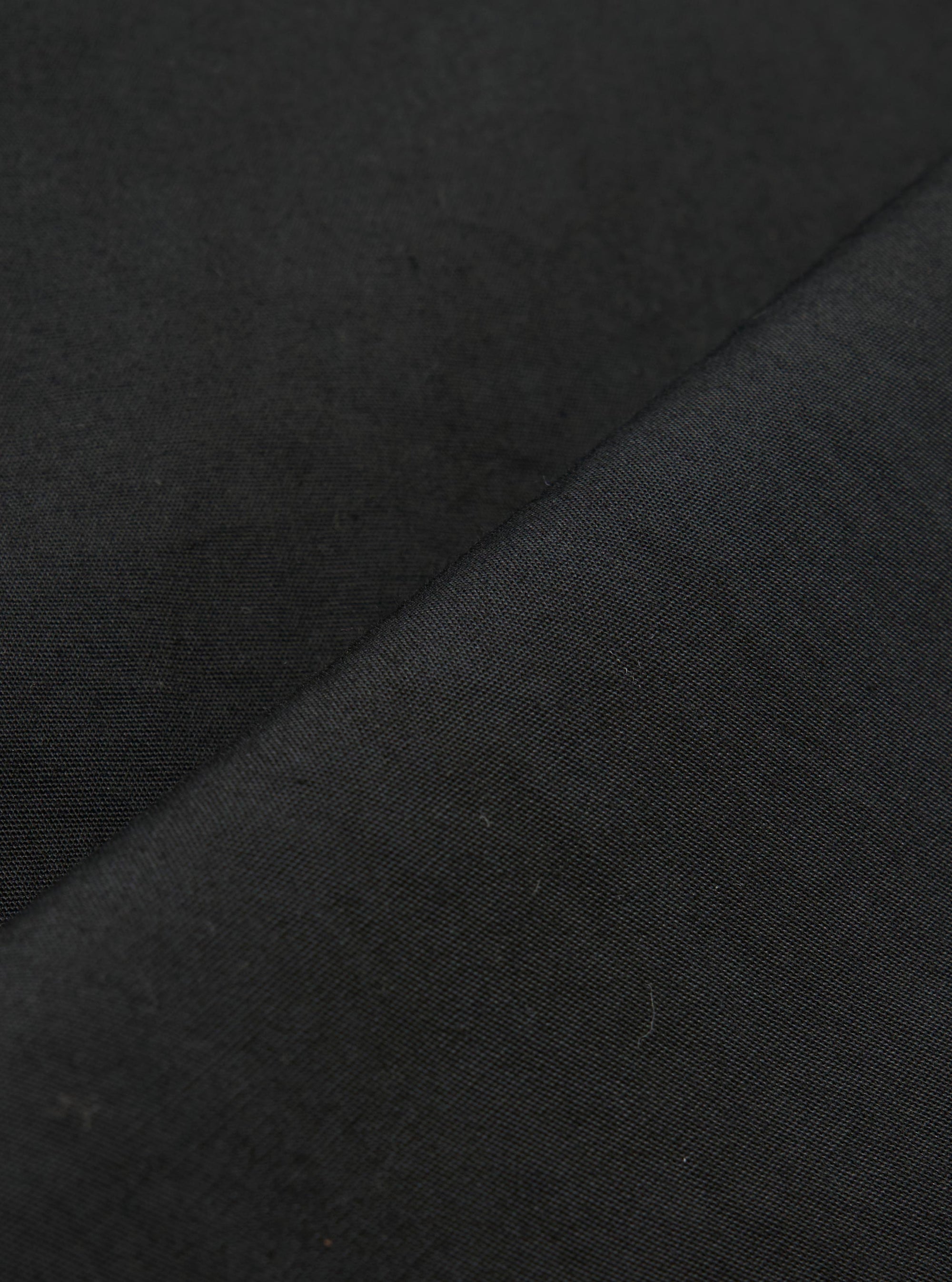 A close-up fabric swatch of the Black Broadcloth Oxford Pant shows a detailed view of the black broadcloth material. The image highlights the smooth, tightly woven texture of the cotton fabric against a neutral background.