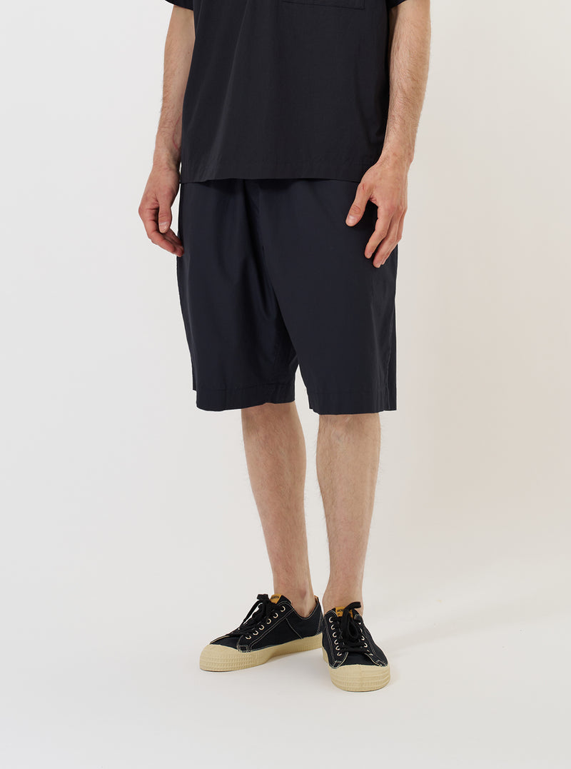 A model stands against a plain white background wearing the Navy Broadcloth Vacation Short. The navy blue shorts feature a relaxed fit and are shown from the waist down, highlighting the smooth texture of the broadcloth fabric and the comfortable, wide-leg cut. The model is pairing the shorts with dark-colored shoes.
