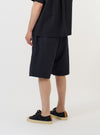 A model is wearing the Navy Broadcloth Vacation Short against a plain white background. The image provides a side-rear view of the shorts, highlighting the relaxed fit, mid-thigh length, and a single rear patch pocket on the right side. The model is standing and pairing the navy blue shorts with dark footwear.